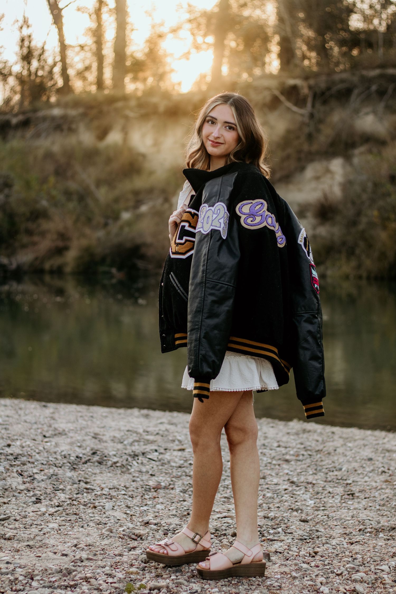 A varsity jacket and boots styled outfit photographed near a lake at sunset with a rocky shoreline.
