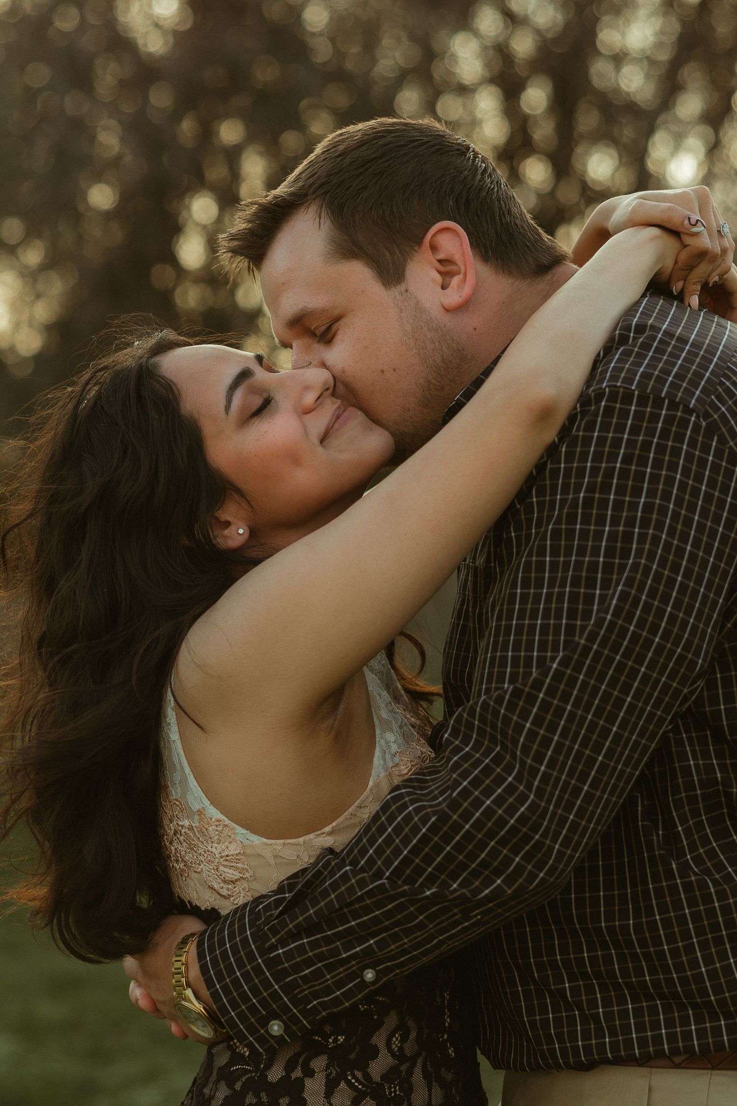A couple shares a romantic embrace outdoors at golden hour, woman smiling as man kisses her cheek.