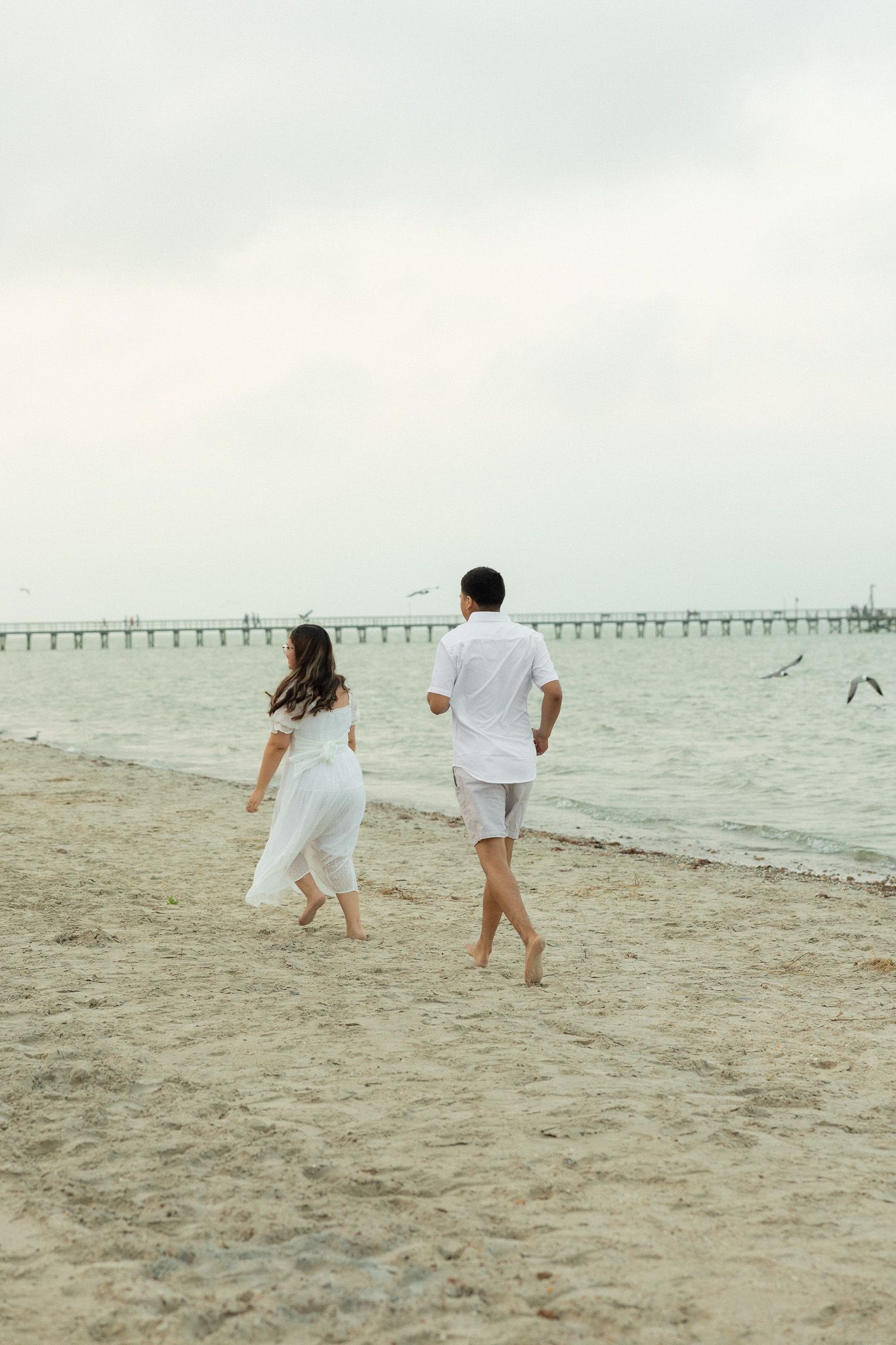 A couple in white outfits walking barefoot on a sandy beach toward the ocean with a pier in the background.