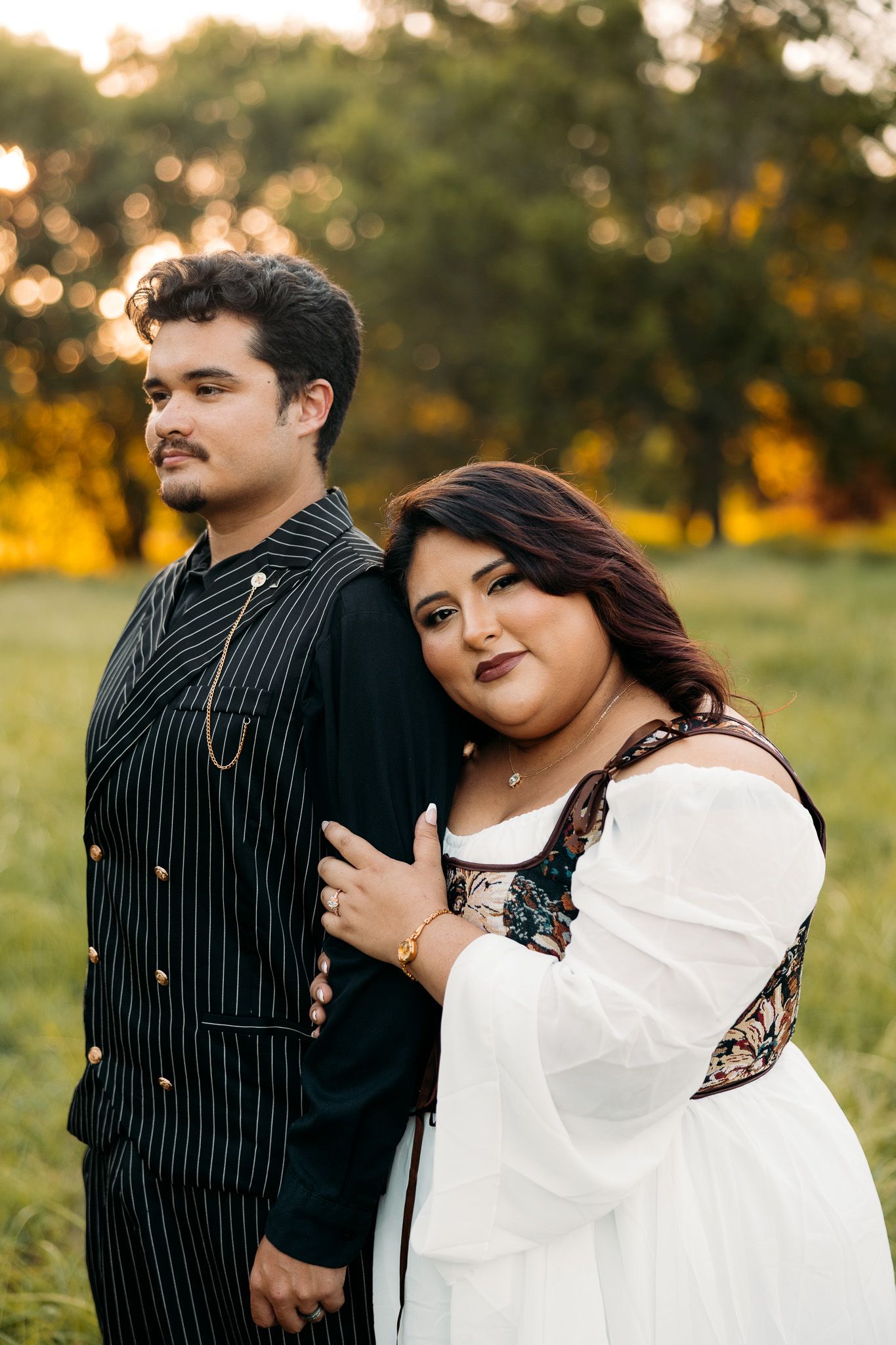 Couple posing outdoors at golden hour, woman in white dress embracing man in black pinstripe suit in a green field.