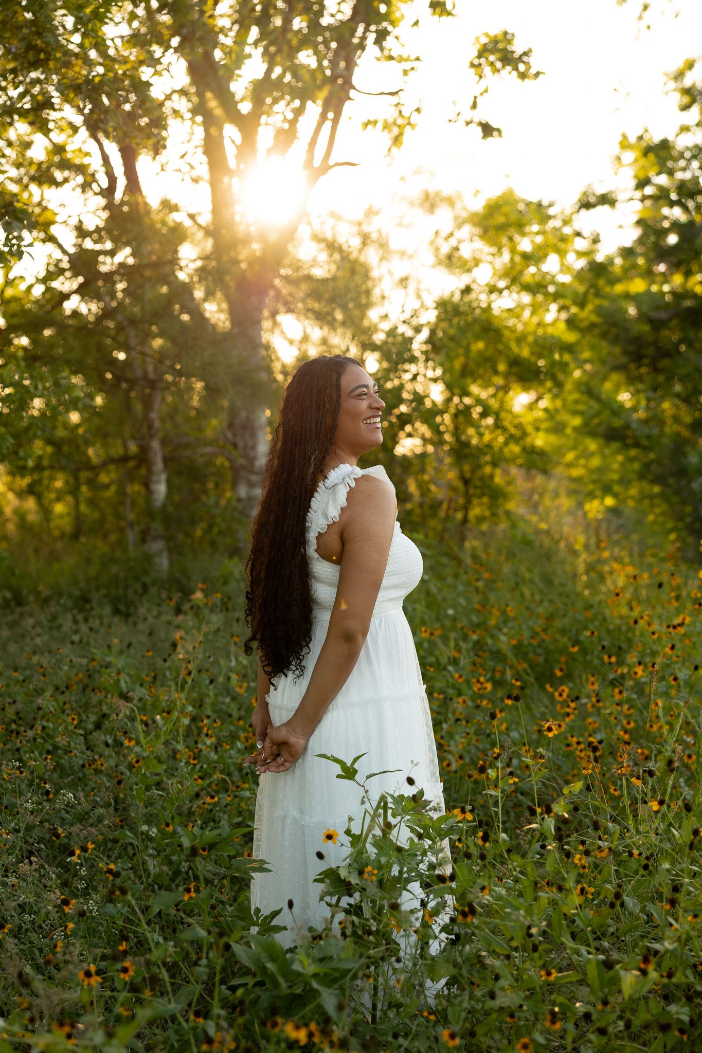Woman in white dress smiling in sunlit meadow with wildflowers and trees at golden hour.
