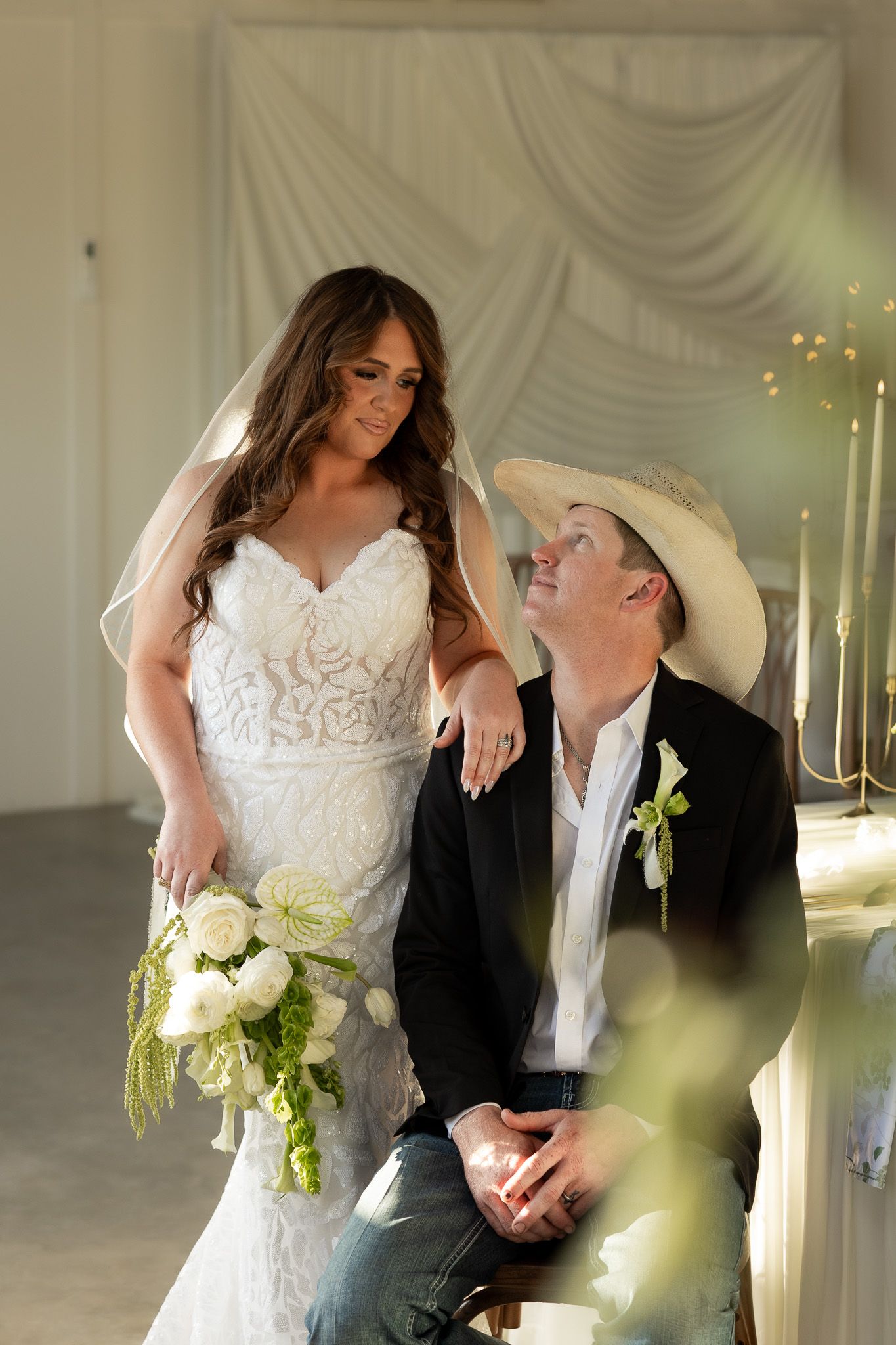 Bride in lace gown holding white bouquet stands beside groom in cowboy hat and black blazer at elegant wedding venue.