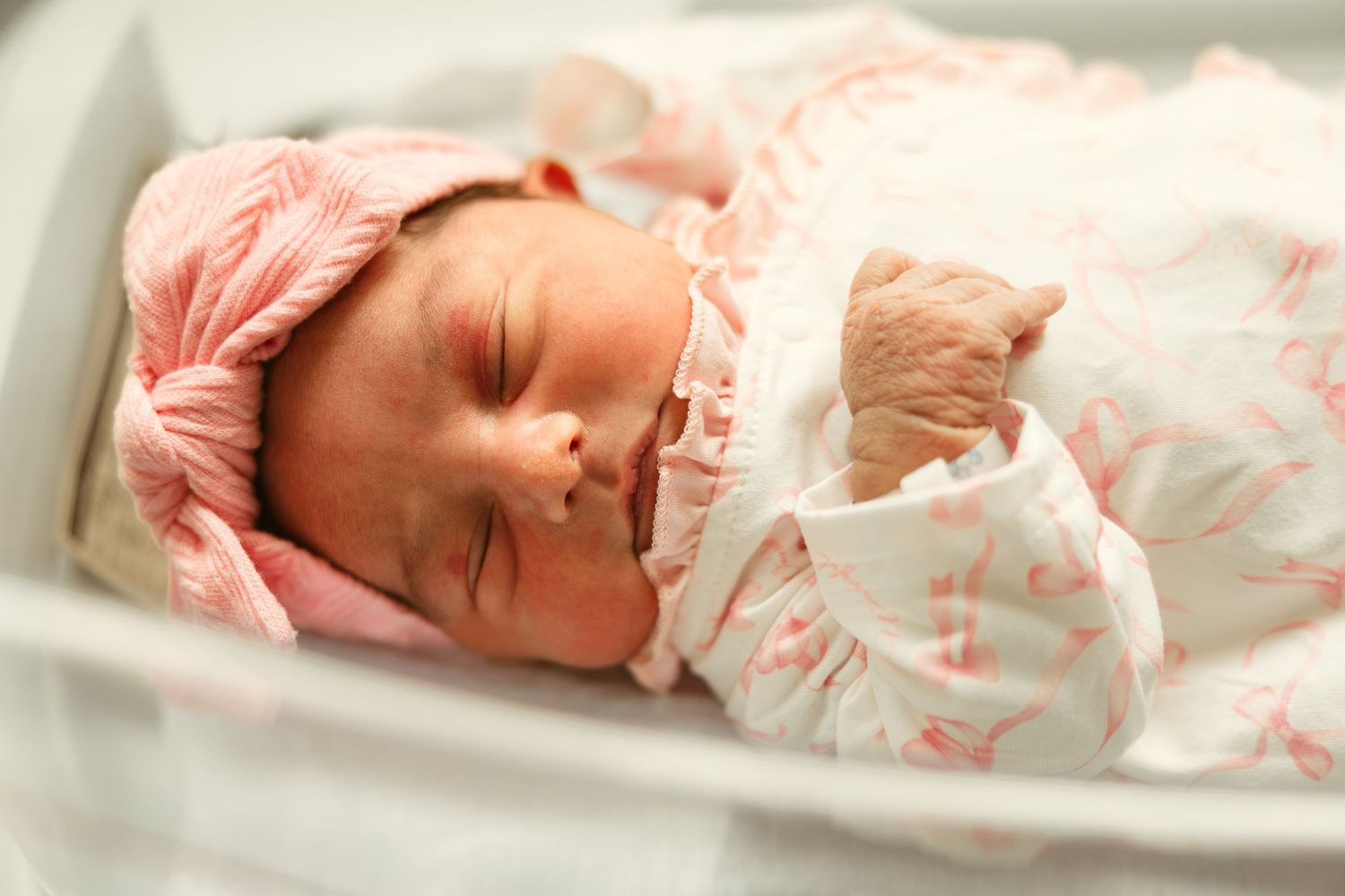 Newborn baby girl sleeping peacefully in hospital bassinet wearing pink floral outfit and matching bow headband.