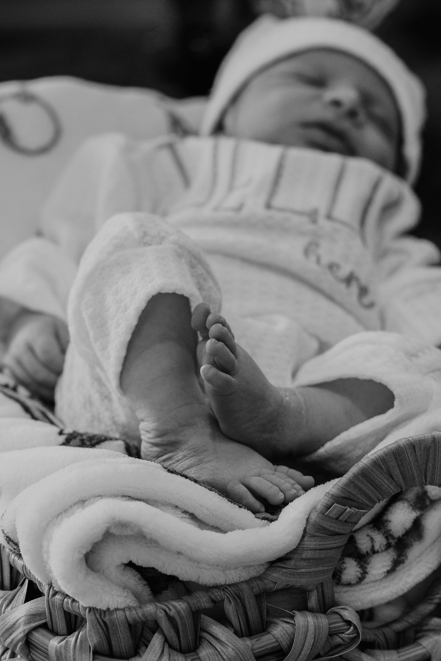 Black and white photo of a sleeping newborn baby in a wicker basket, wearing a white onesie and hat.