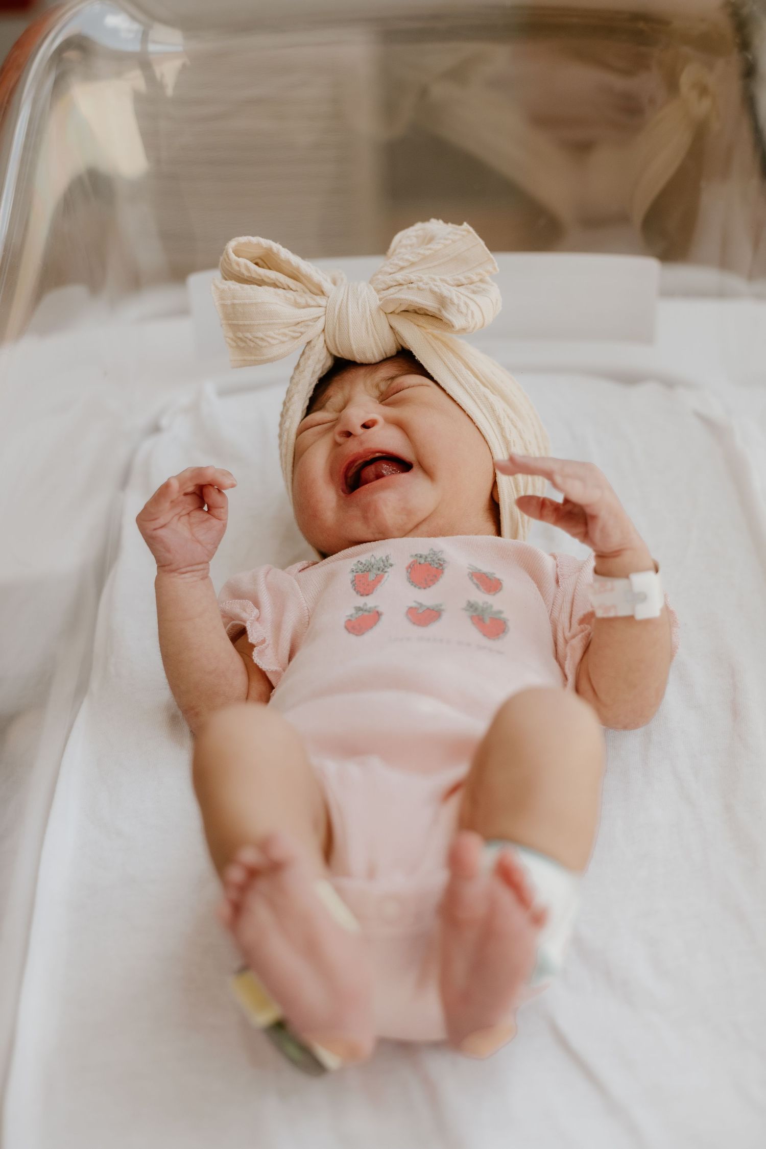 Crying newborn baby girl in pink strawberry onesie and bow headband lying in hospital bassinet.