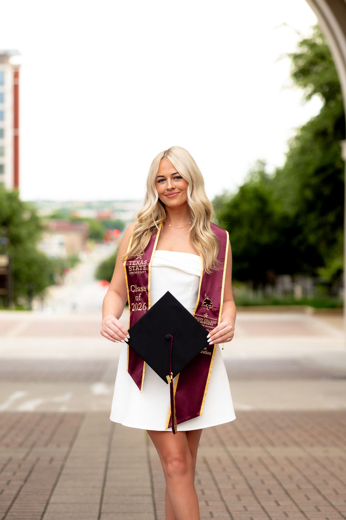 Smiling blonde graduate in white dress holding cap, wearing maroon Class of 2024 sash, posing on urban street.