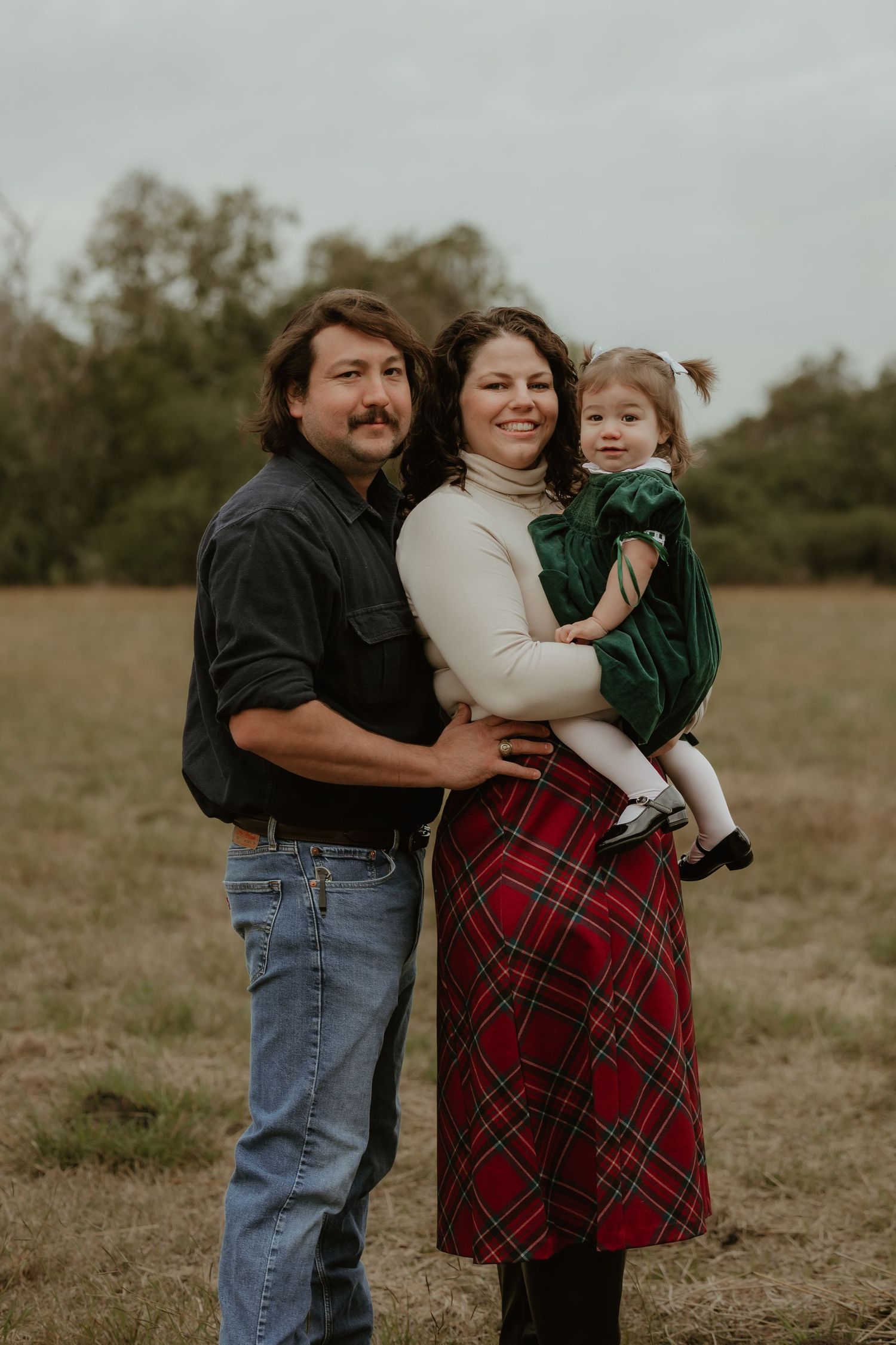 Family of three posing in a field, parents holding toddler in green dress, woman in red plaid skirt, man in dark shirt.