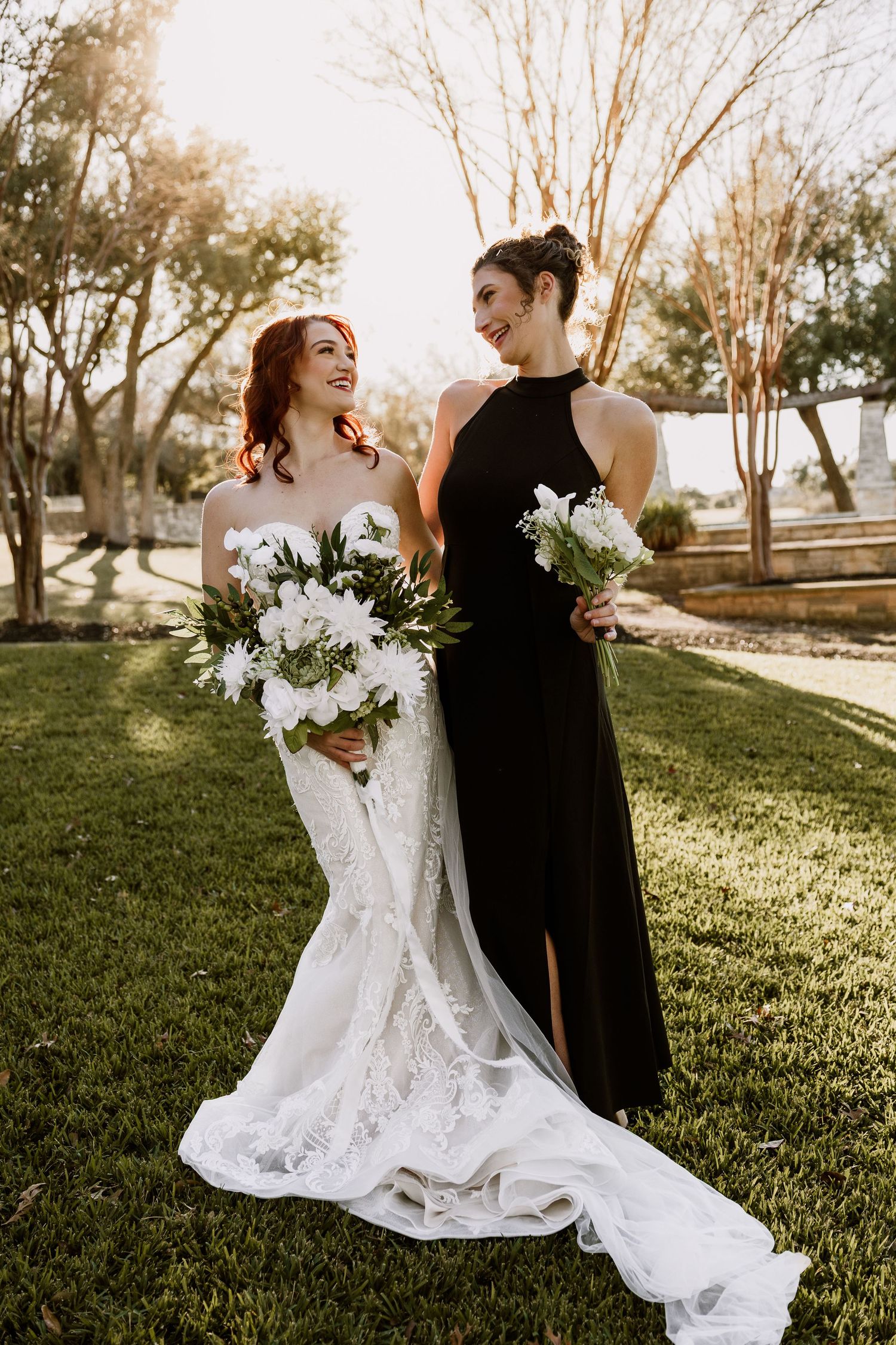 A bride in a flowing white lace gown twirls joyfully with someone in black against a sunlit grassy backdrop.