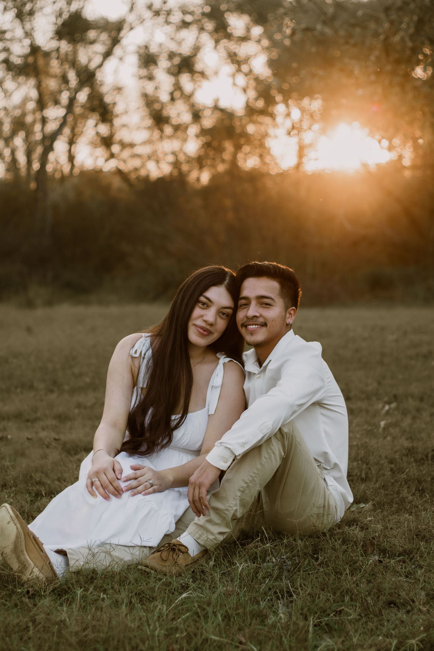 A couple in white clothing lies together on grass during golden hour sunset.