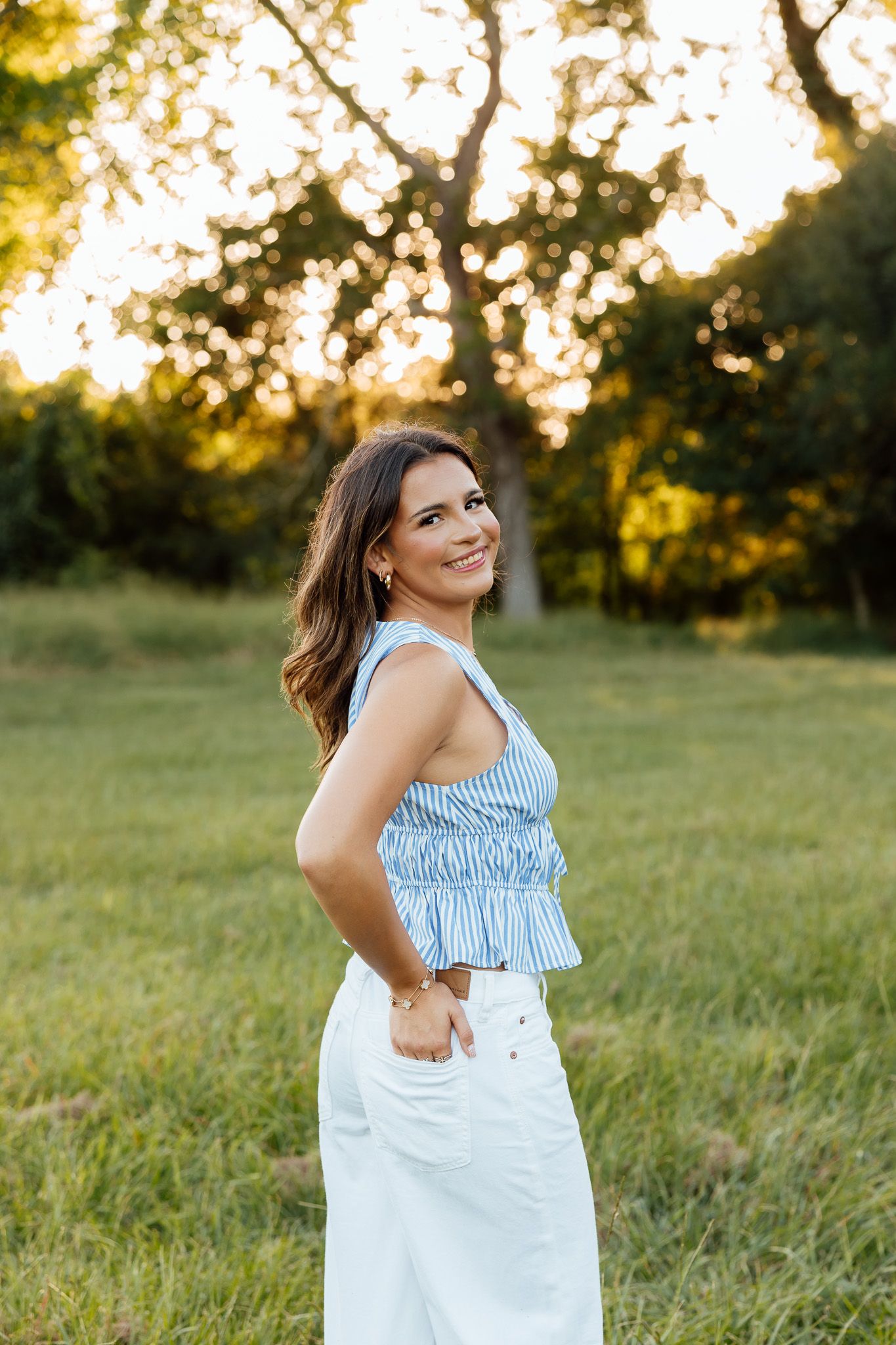 Young woman in blue striped top and white skirt smiling outdoors at golden hour sunset.