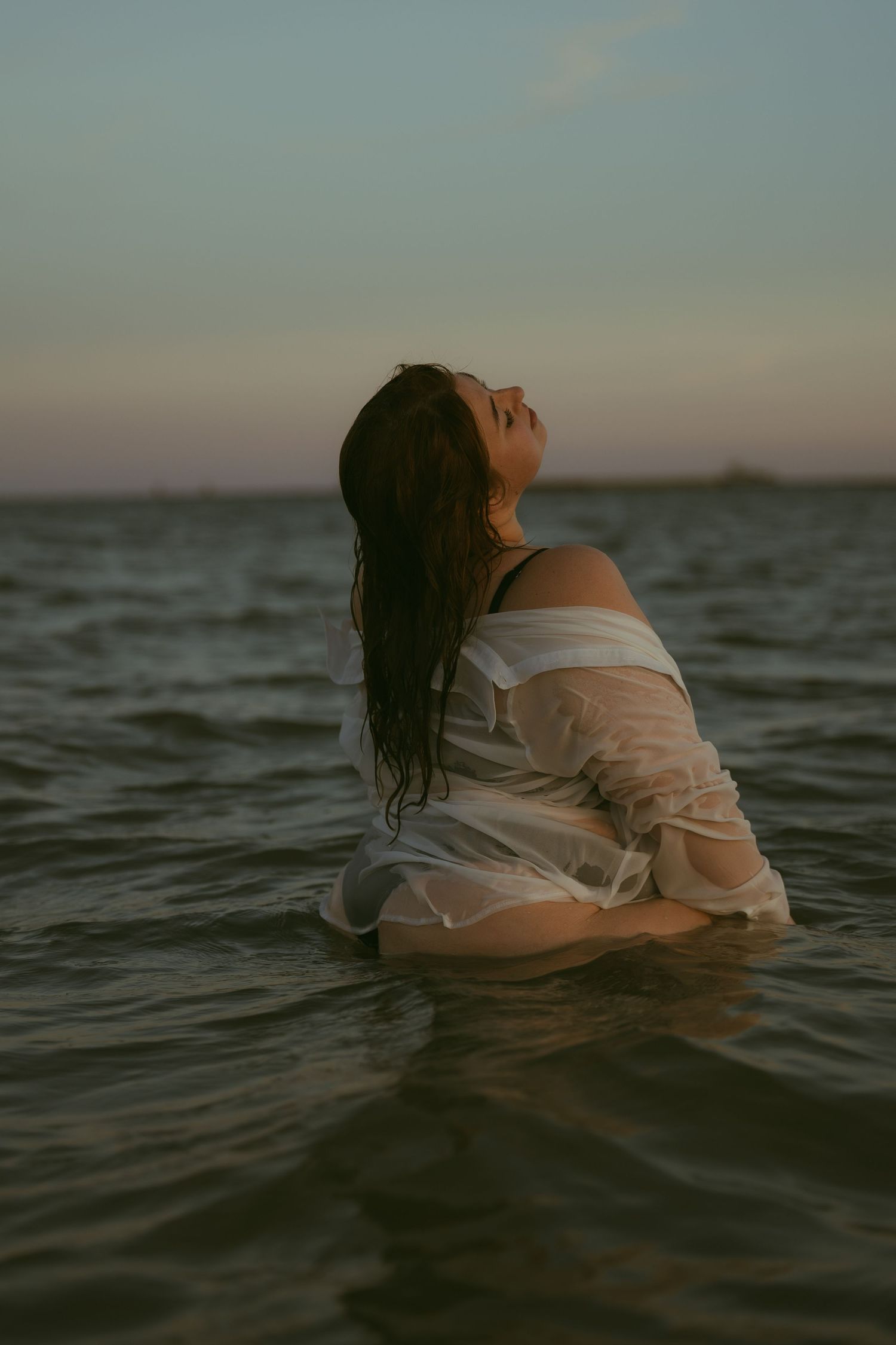 Woman in wet white sheer shirt kneeling in ocean water at dusk, head tilted back with long dark wet hair.