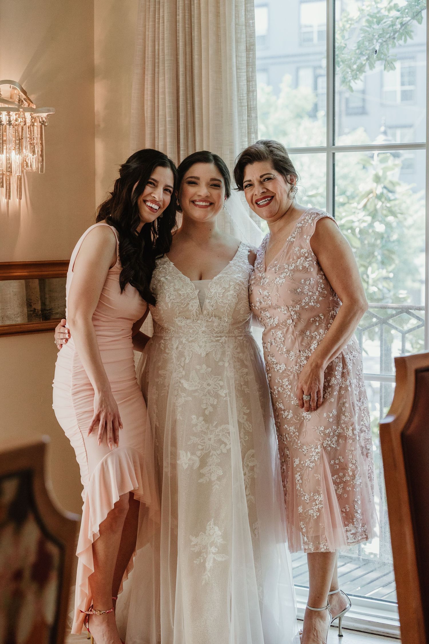 Three women in formal dresses pose together by a window in celebration.