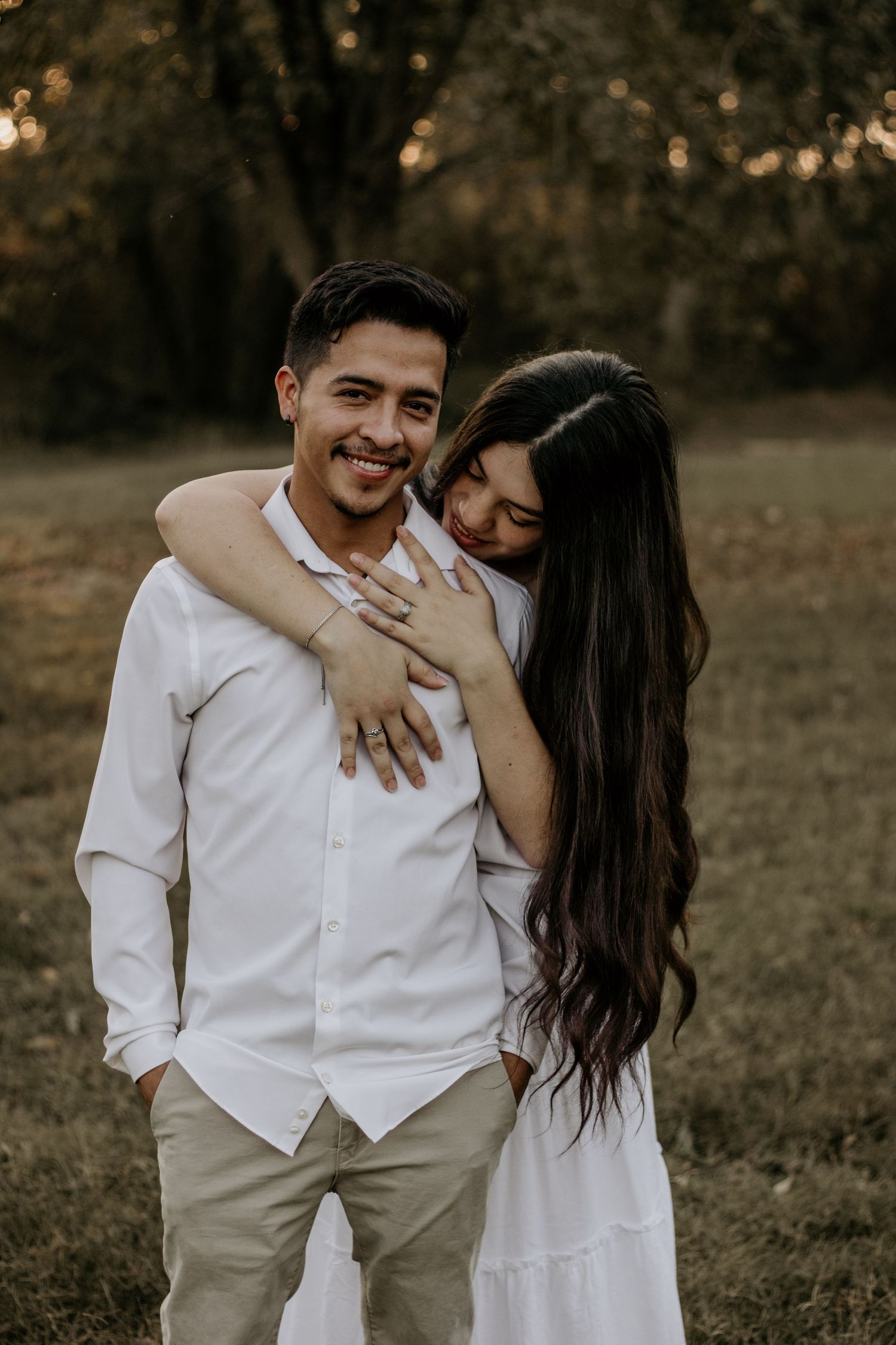 A romantic couple in white outfits embraces and laughs together during a sunset photoshoot in a grassy field.