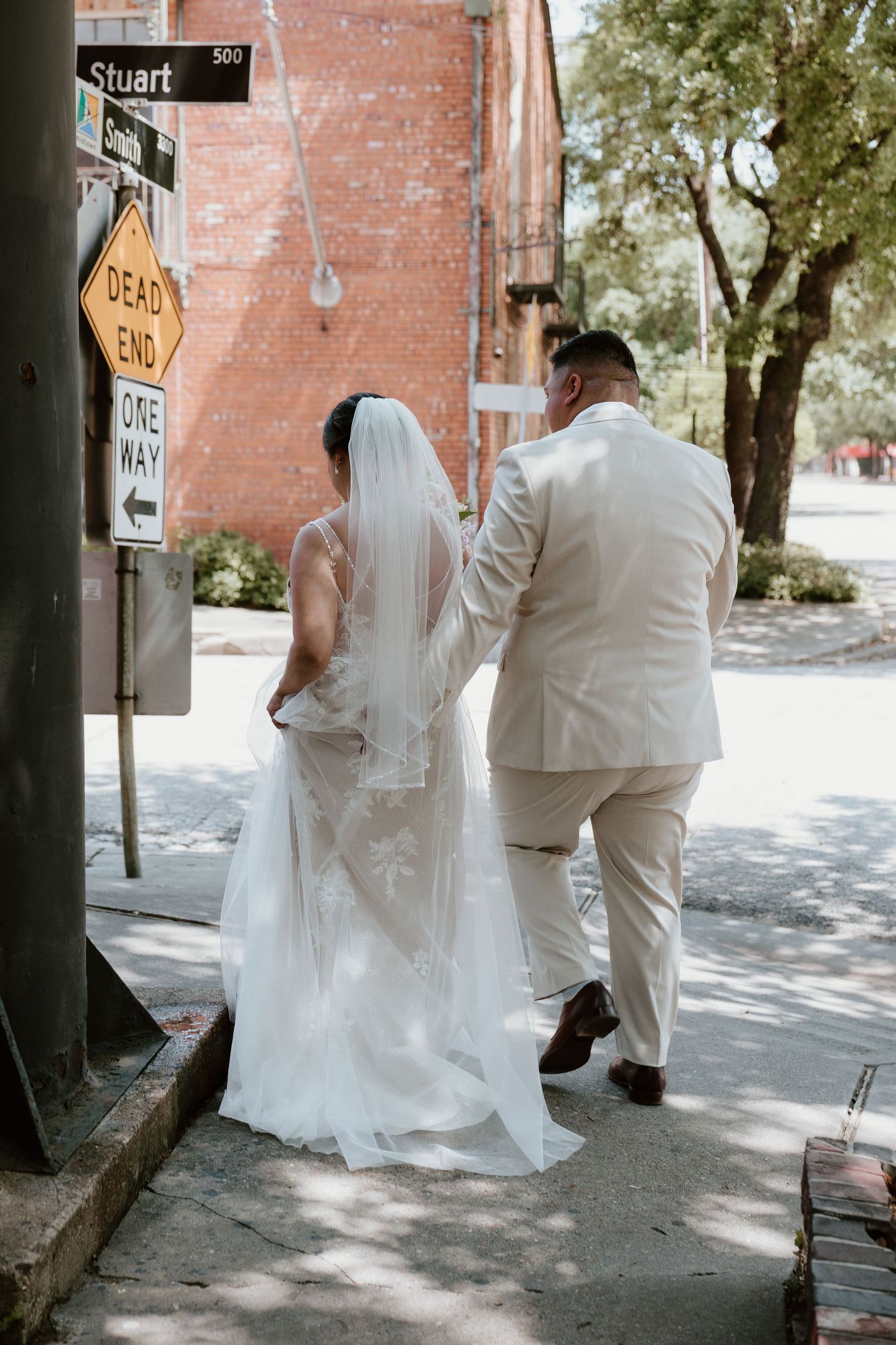 A wedding couple stands near a street sign in their elegant white and cream attire.