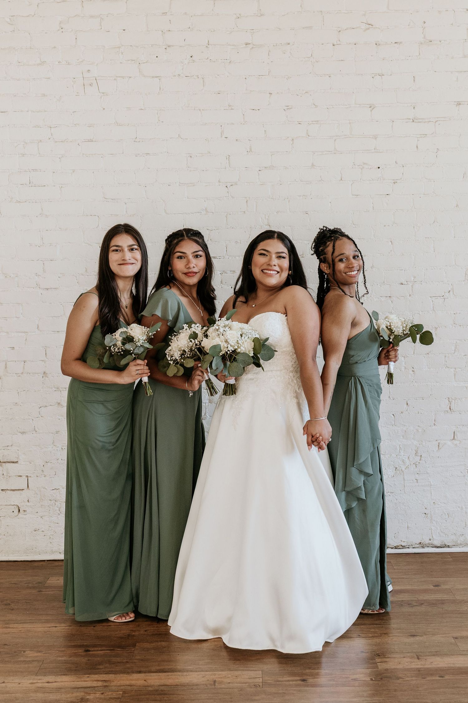 Bridesmaids in sage green dresses pose with bride in white gown against brick wall.