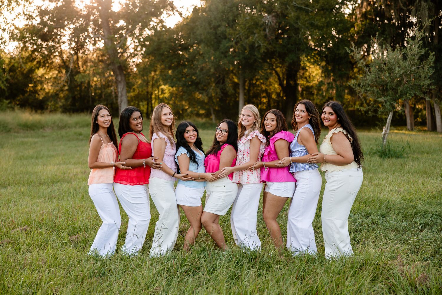Eight women in white pants pose in a line outdoors at golden hour in a lush green field.