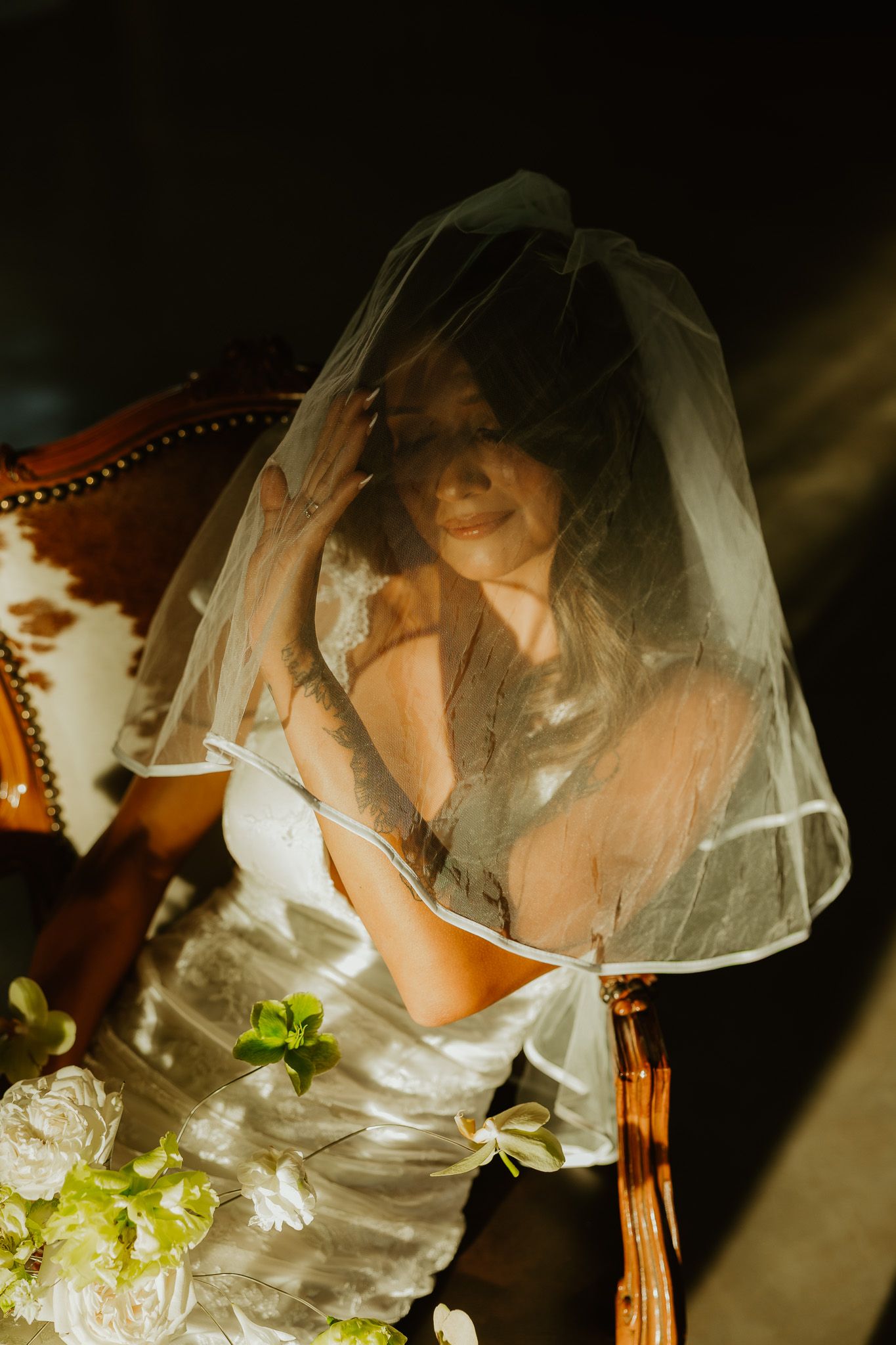 Bride in white gown and veil seated in ornate chair with soft sunlight and white floral arrangement.