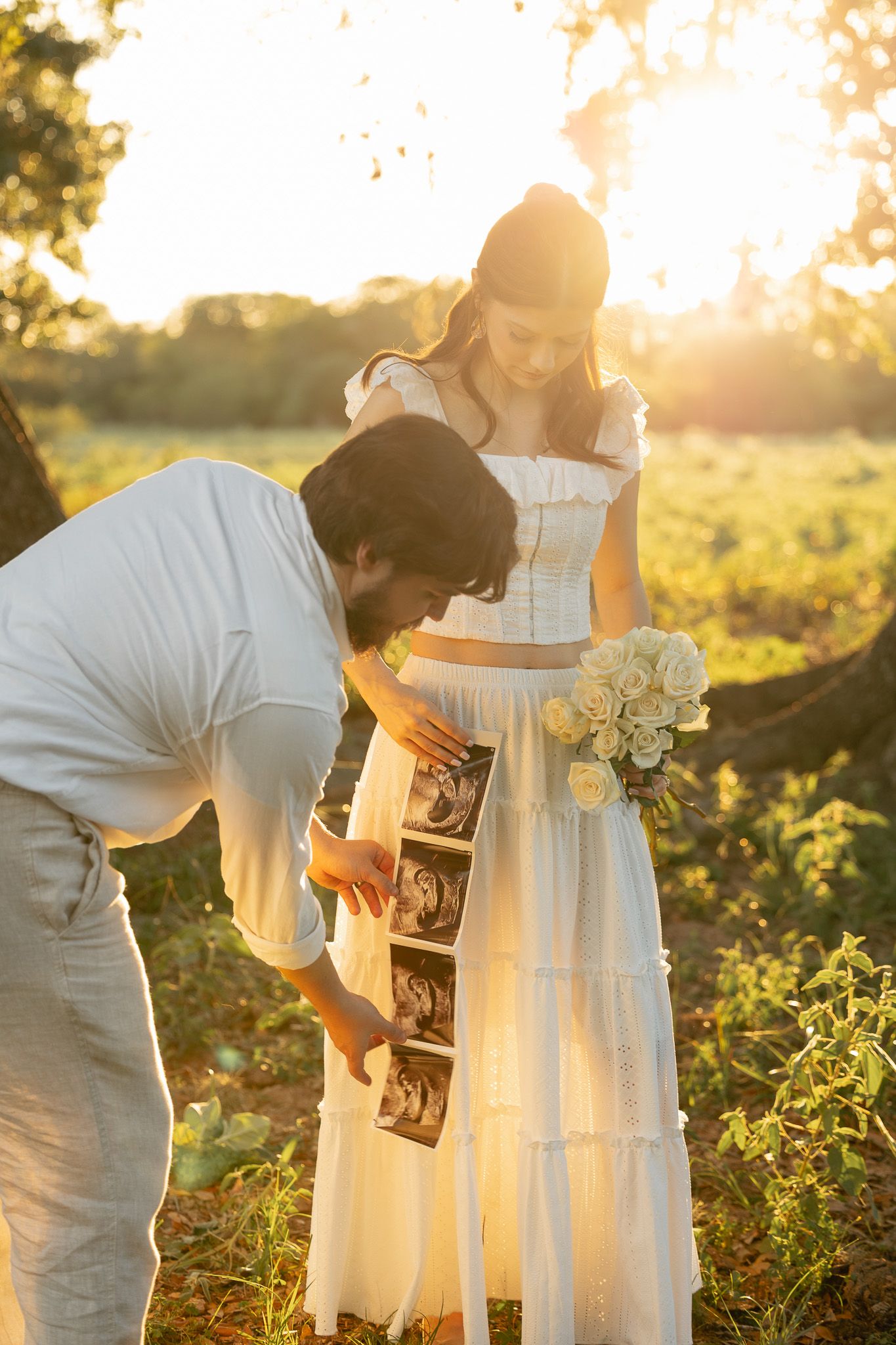 Couple in white outfits examining ultrasound photos outdoors at golden hour, pregnant woman holding white roses bouquet.