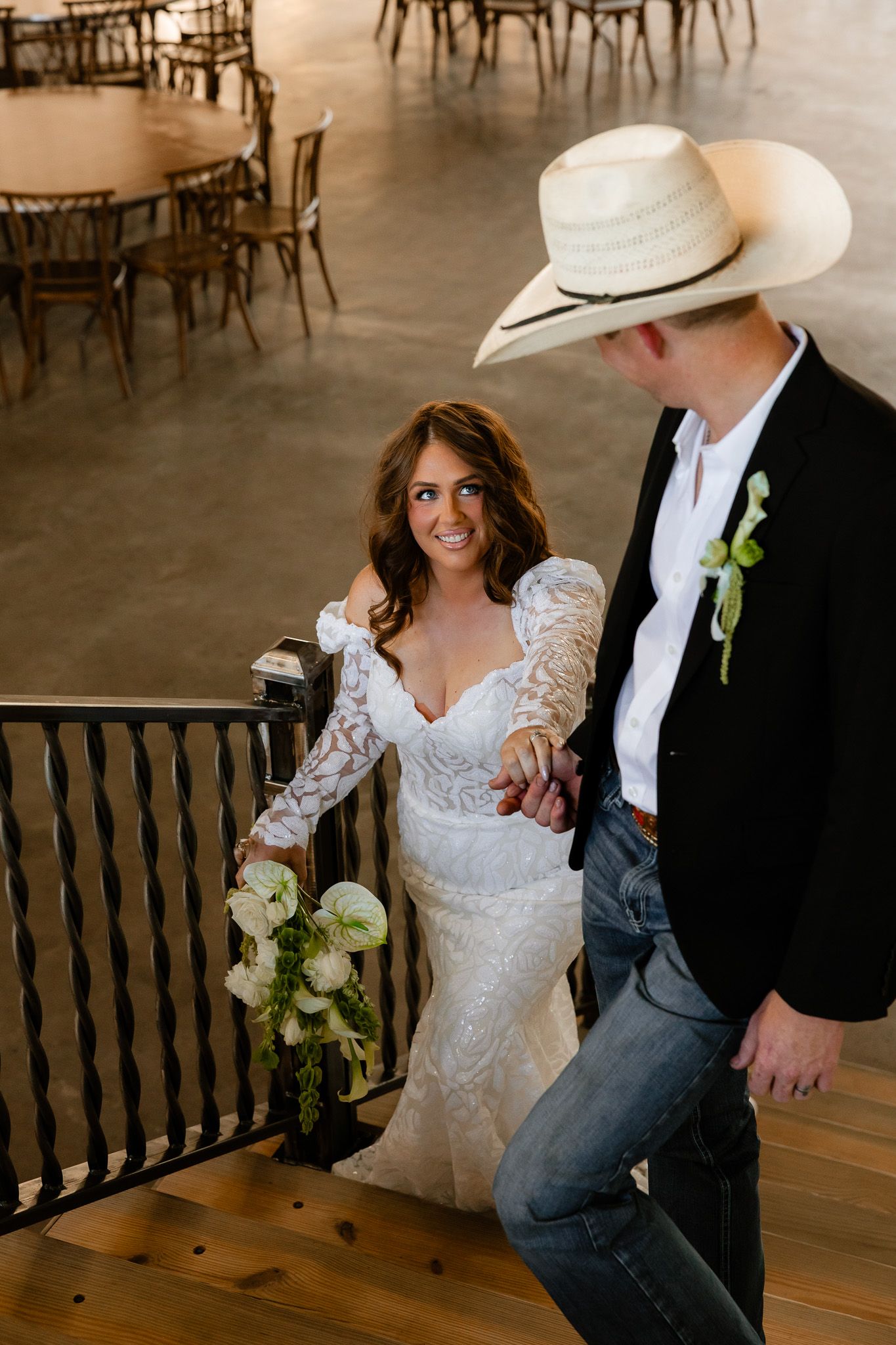 Bride in lace gown holds bouquet while groom in cowboy hat and jeans leads her up wooden stairs at rustic wedding venue.