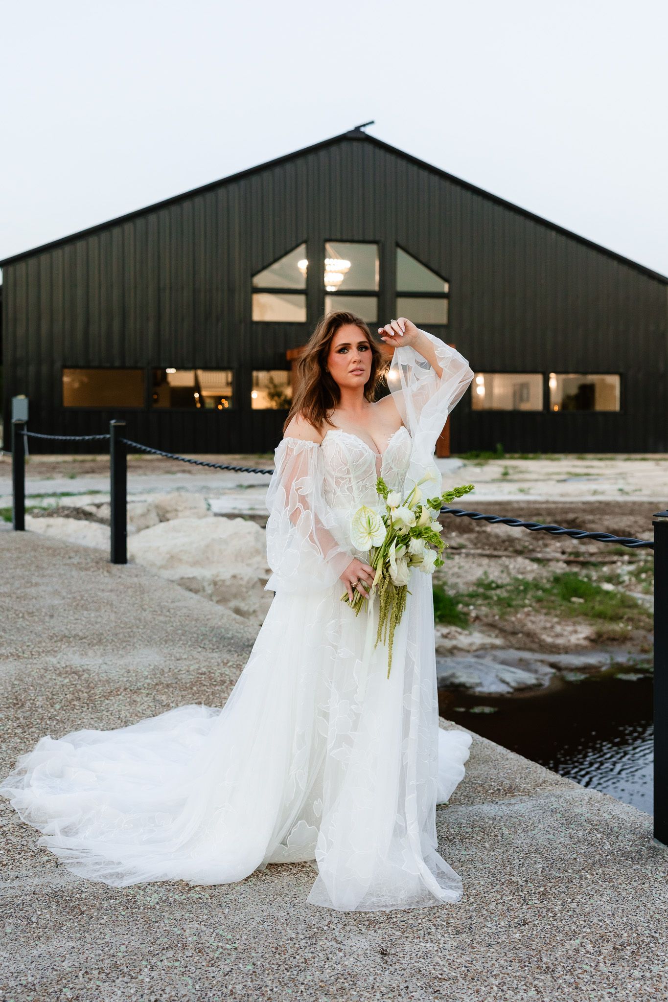 Bride in white lace gown with sheer sleeves holds bouquet outside black barn venue.