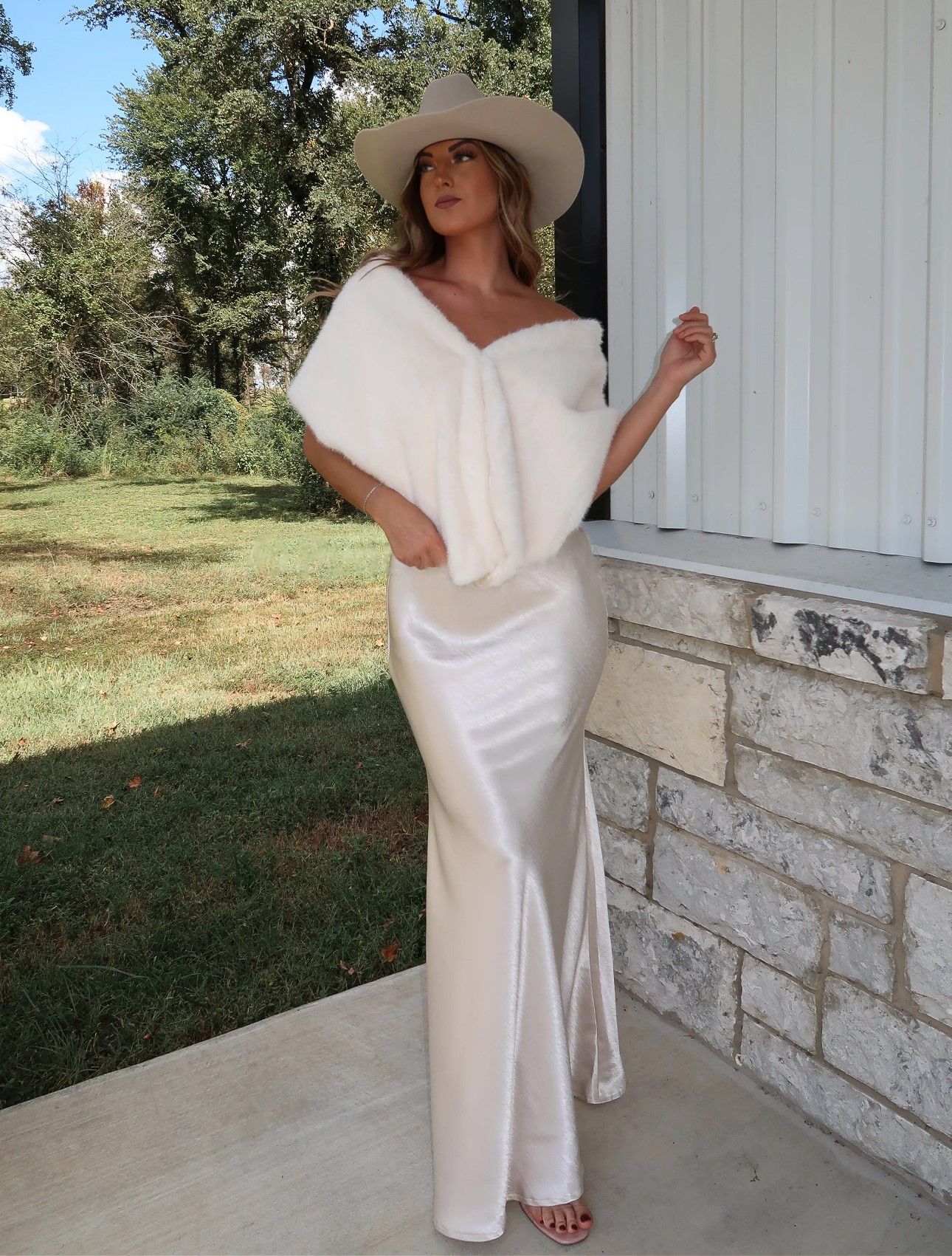 A figure in a flowing white dress and wide-brimmed hat poses elegantly on a stone porch in a rustic outdoor setting.