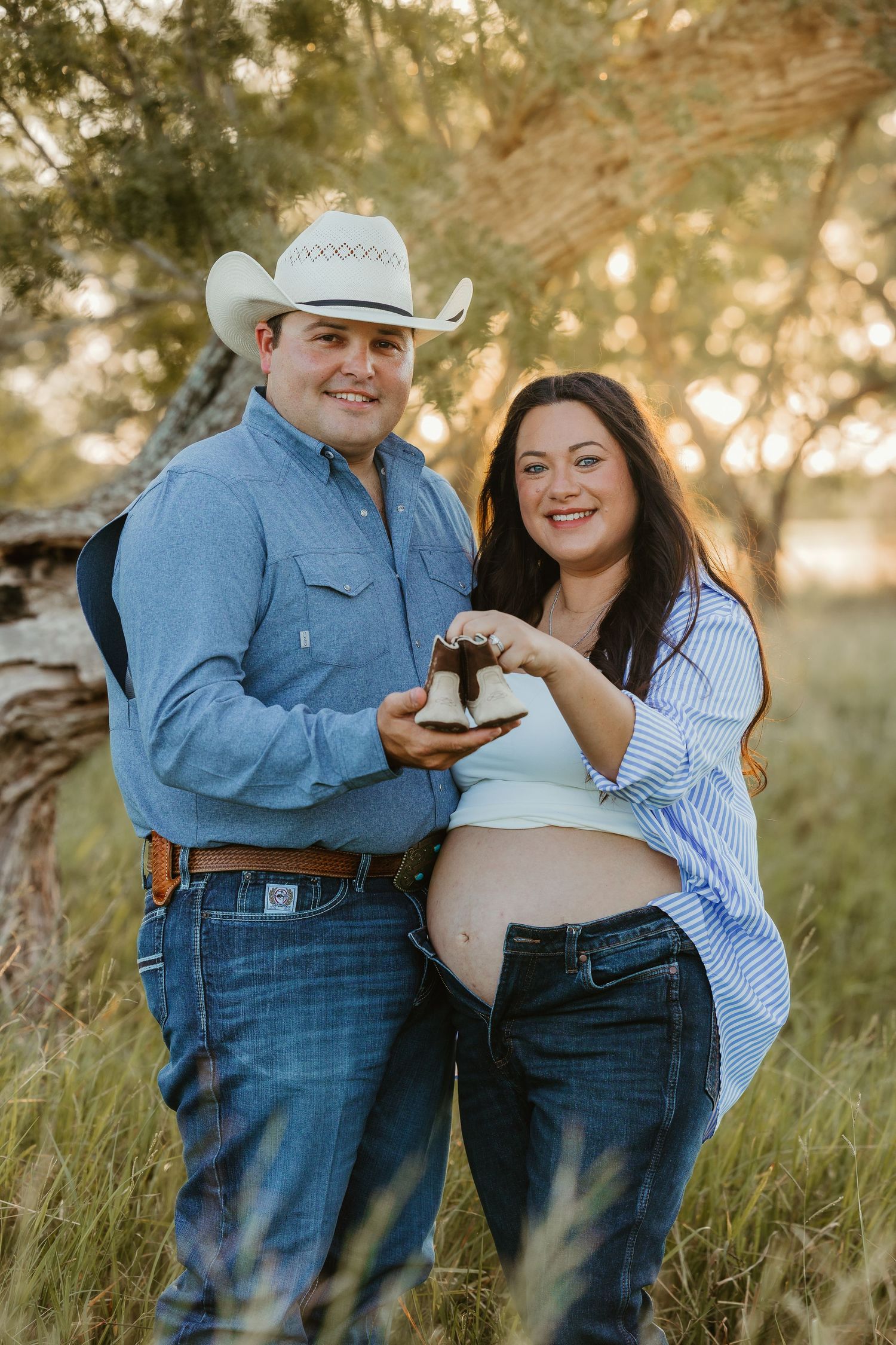 An expectant couple shares a tender moment while holding baby shoes in a field.
