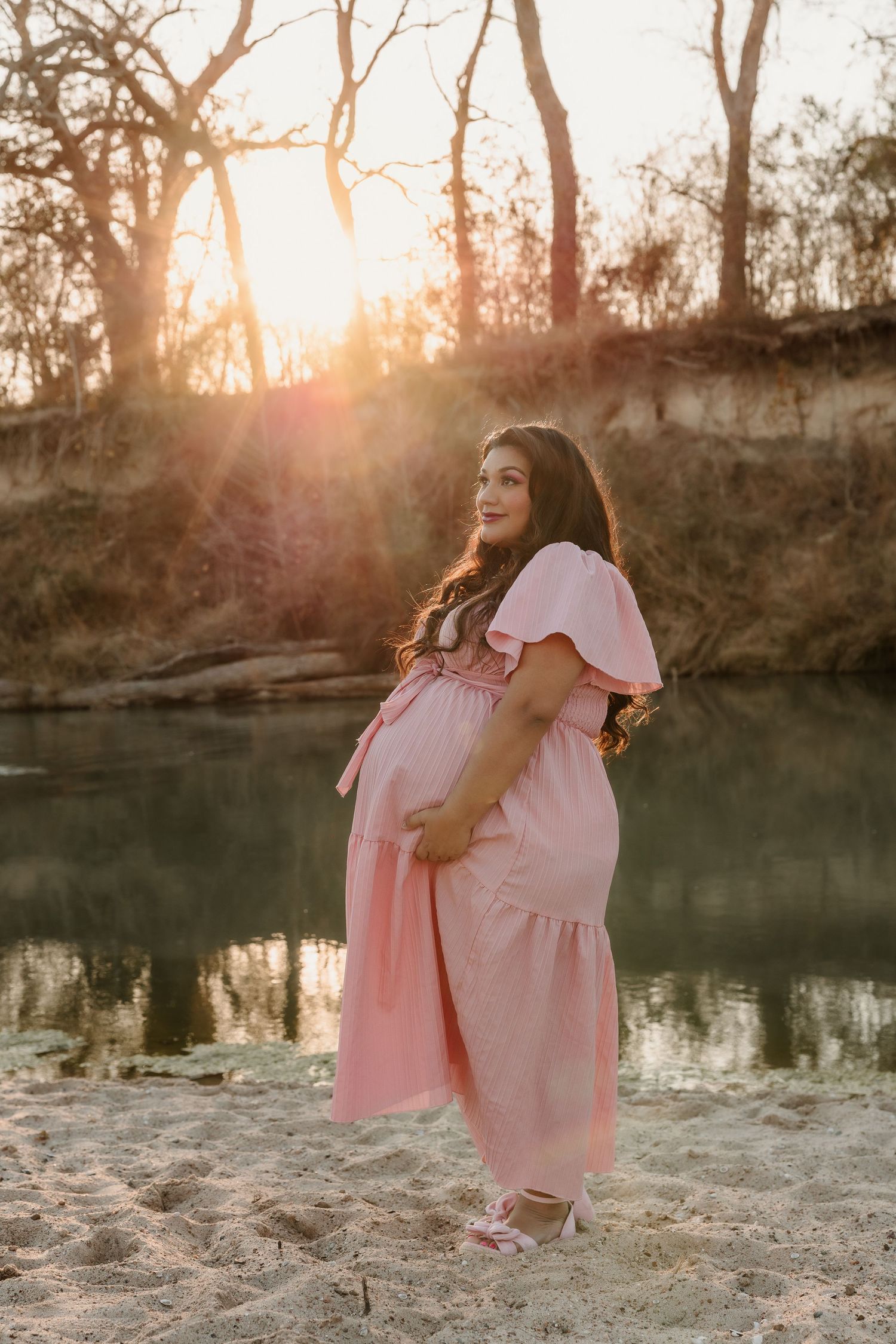 Pregnant woman in pink maxi dress poses by a river at golden hour sunset on sandy shore.