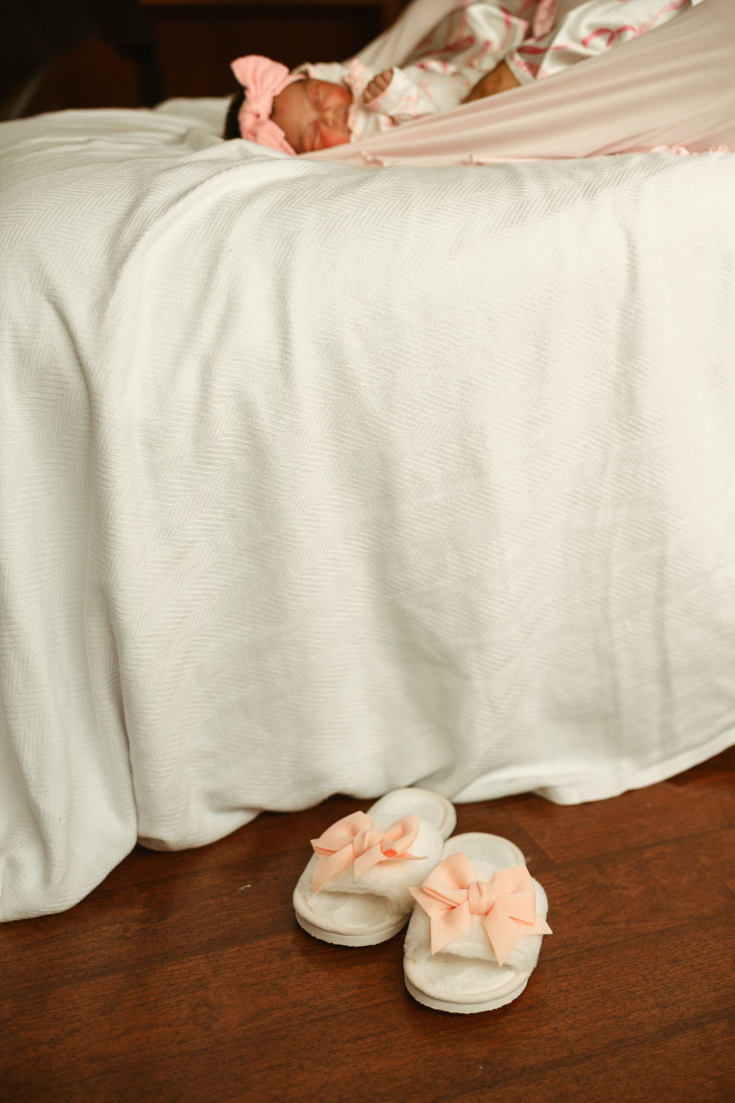 Newborn baby resting on a white bed with pink bow slippers placed on the wooden floor in the foreground.