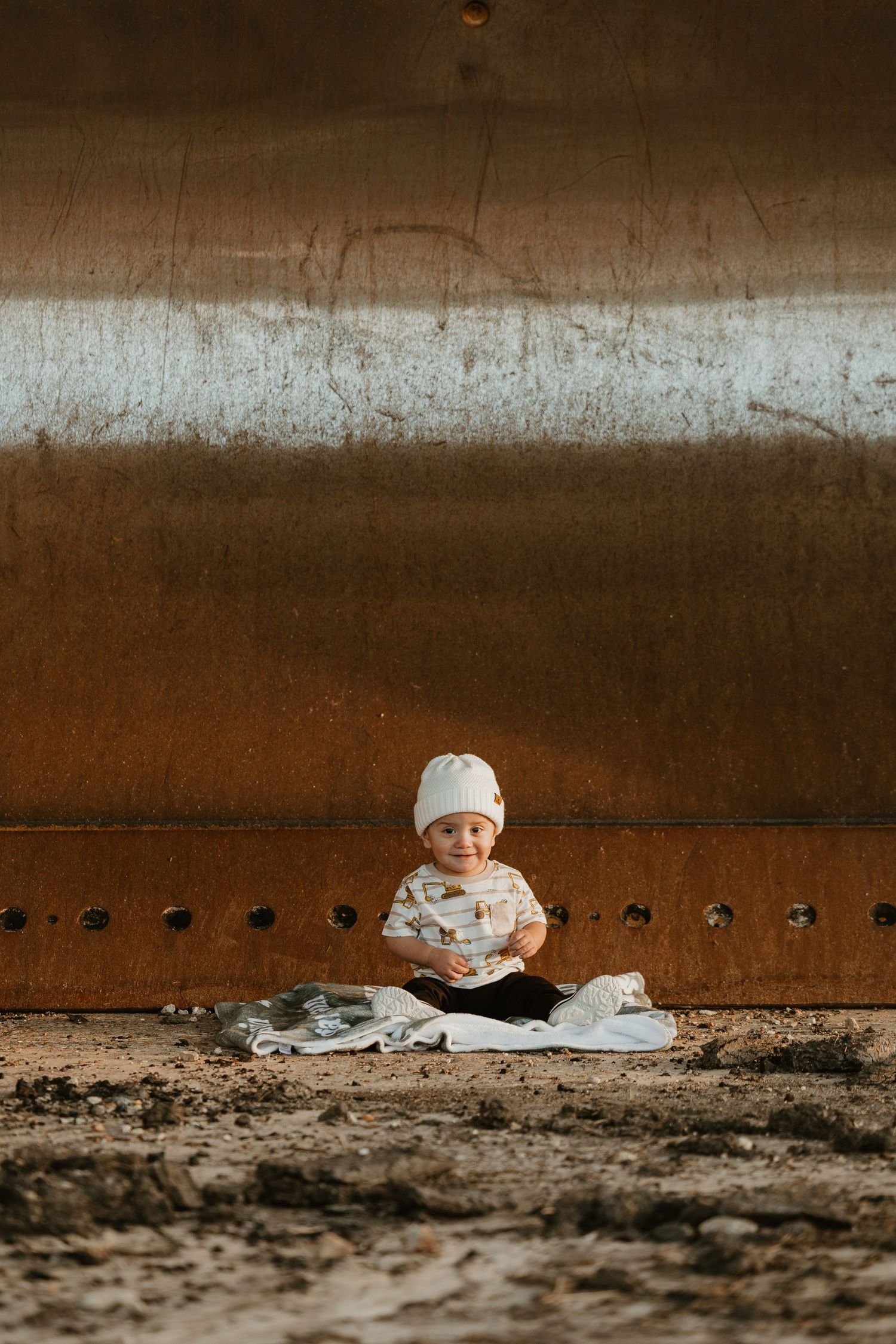 A smiling baby in a white beanie sits on sandy ground against a rusty metal wall outdoors.