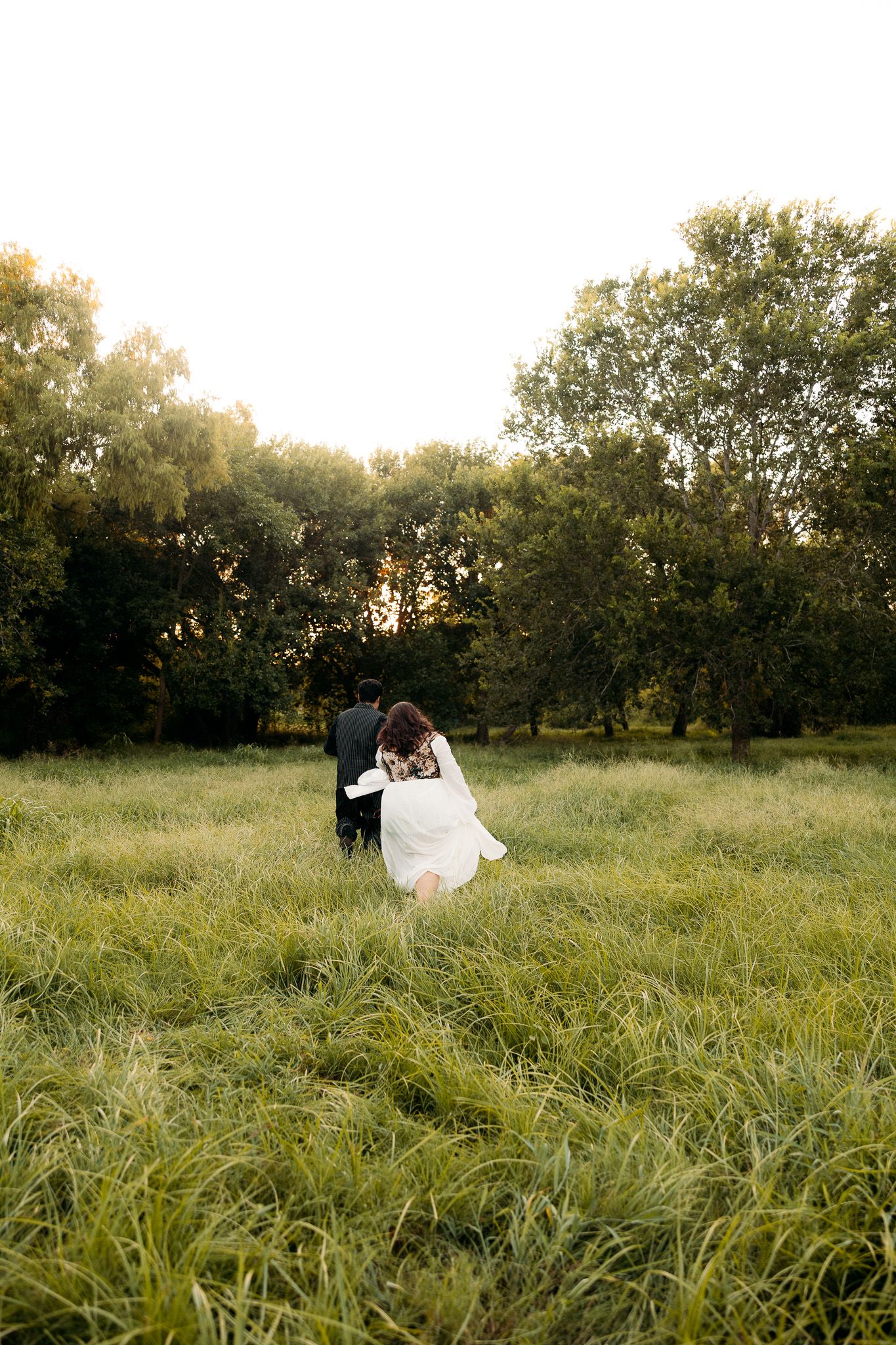 A bride and groom walk hand in hand through a lush green meadow at golden hour with trees in the background.
