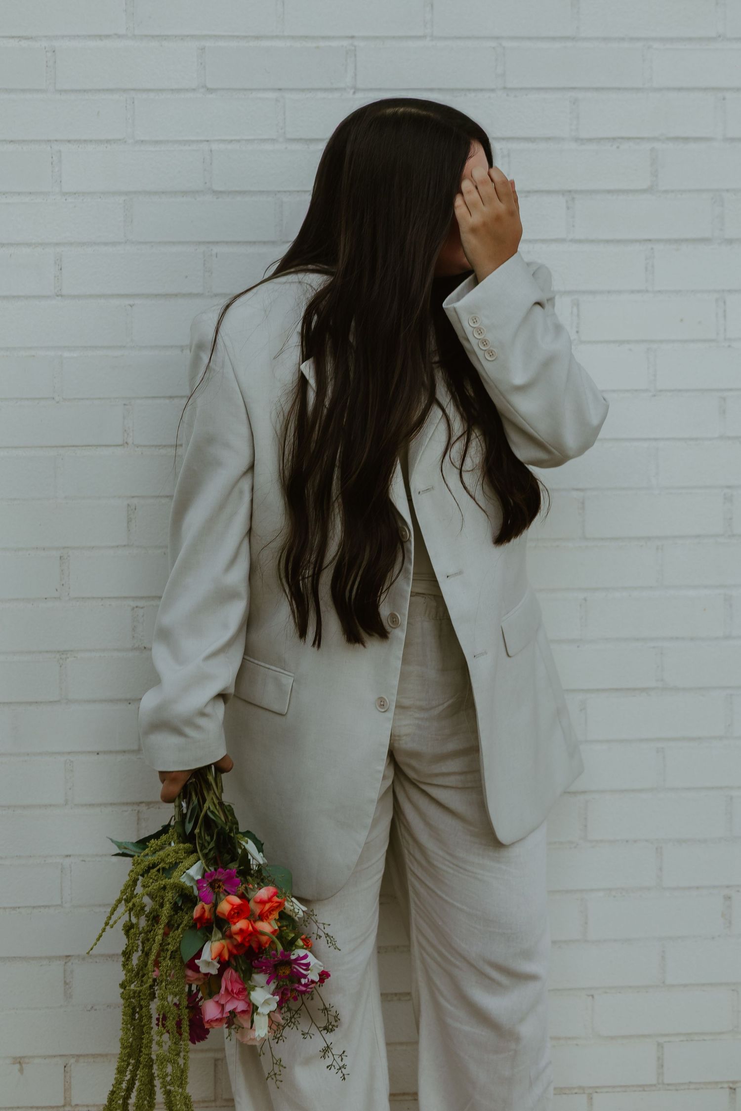 Artistic portrait against white brick wall showing person in light suit holding bright flowers.