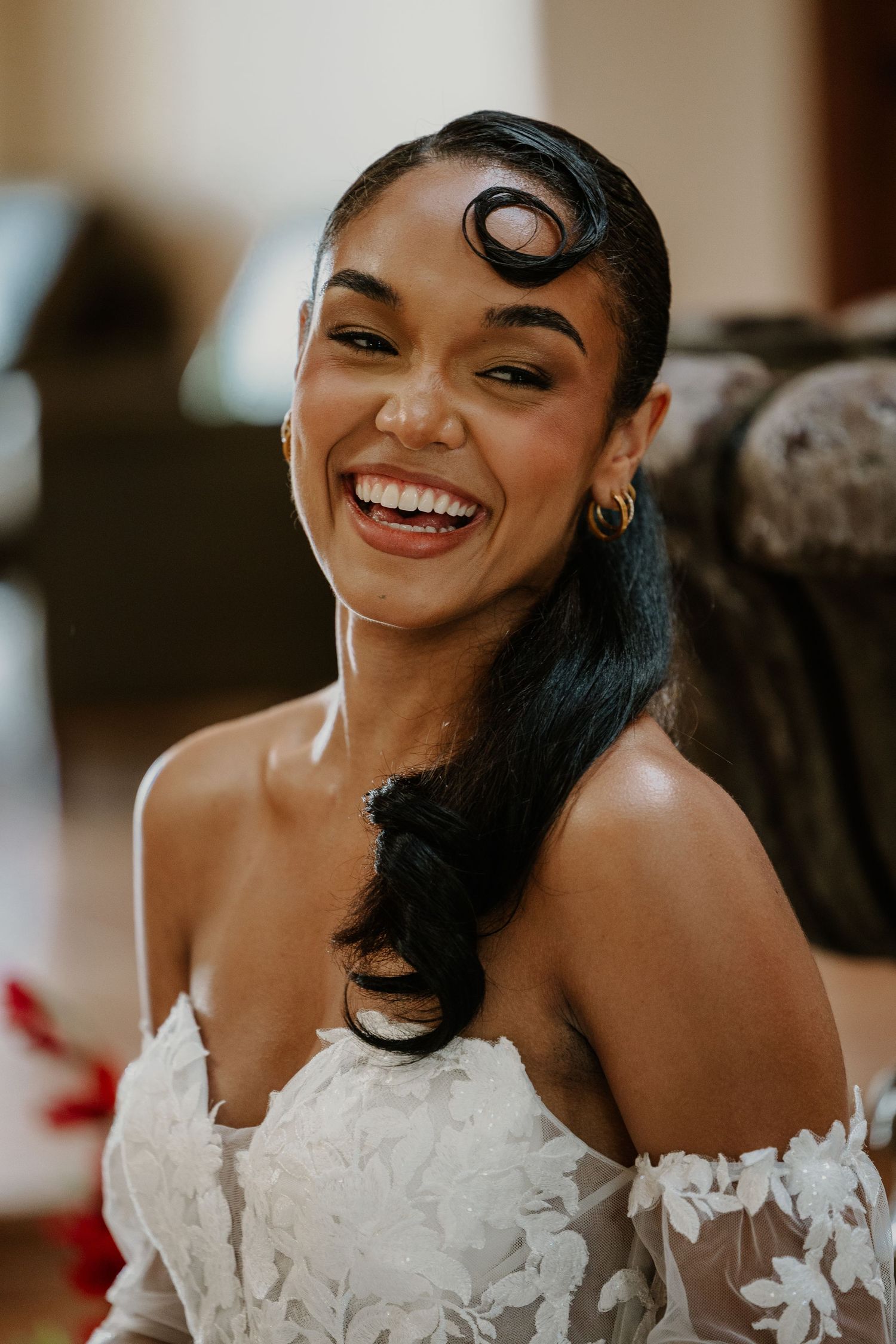 A bride wearing an off-shoulder lace wedding dress smiles joyfully with vintage-inspired finger waves in her dark hair.