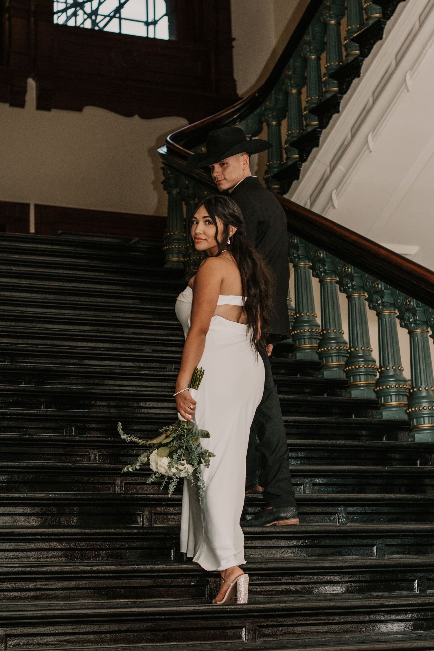 A couple poses on grand dark stairs in formal attire during an indoor photoshoot.