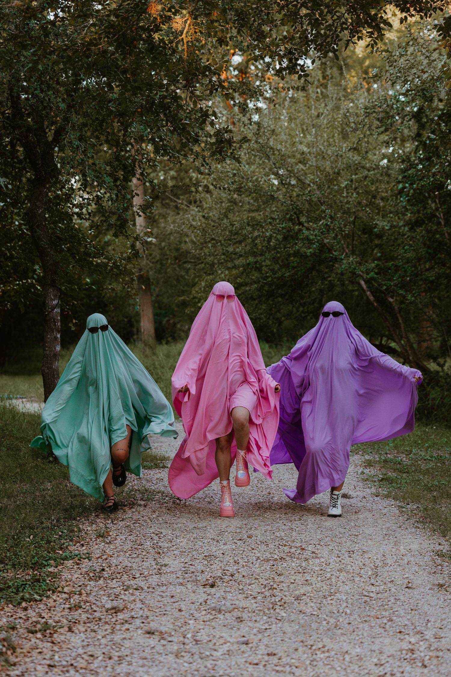 Row of ghostly figures in mint, pink and purple dresses walking along woodland path.