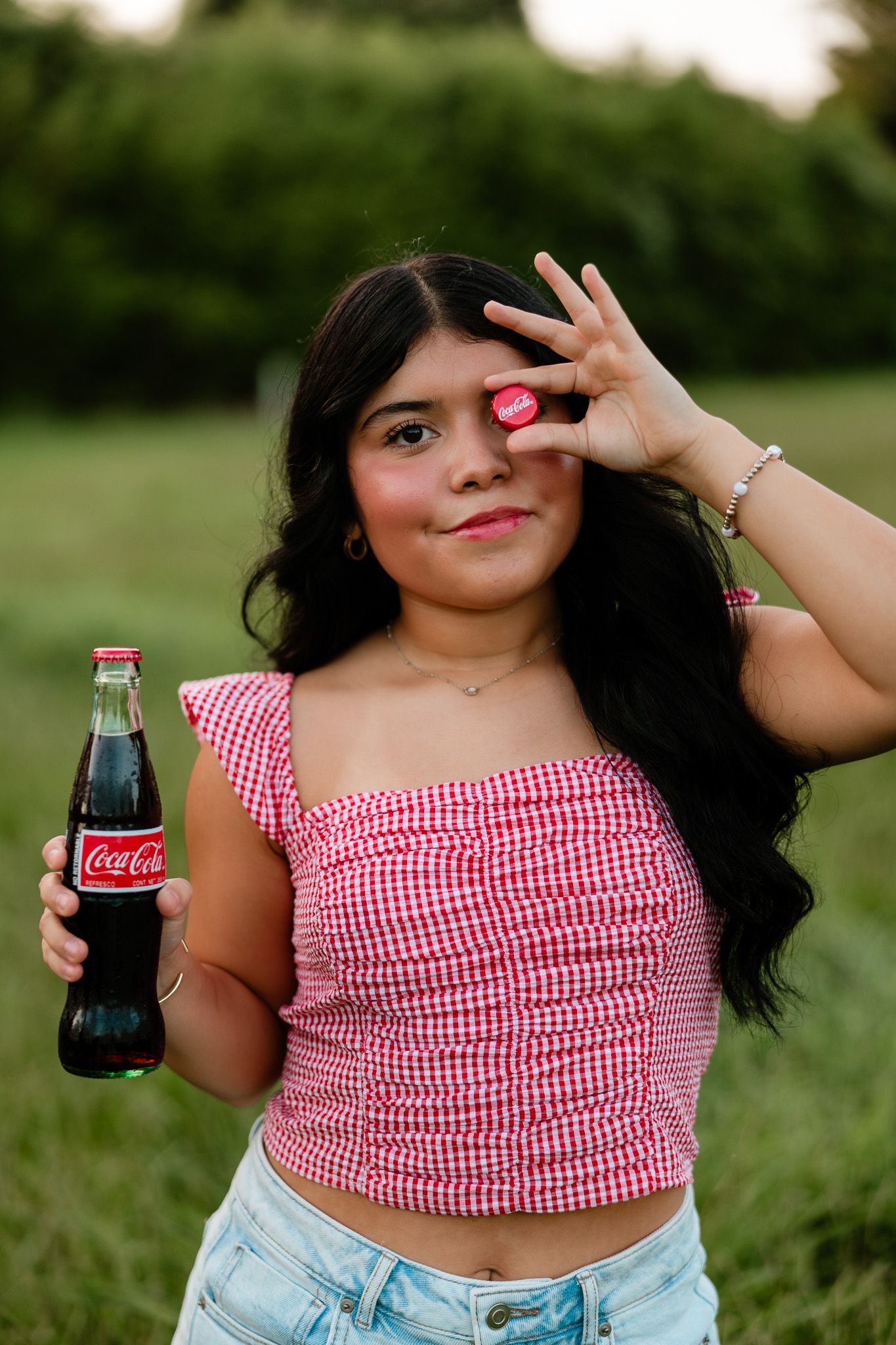 Young woman holding a Coca-Cola glass bottle outdoors, wearing a red gingham crop top and jeans, smiling at camera.