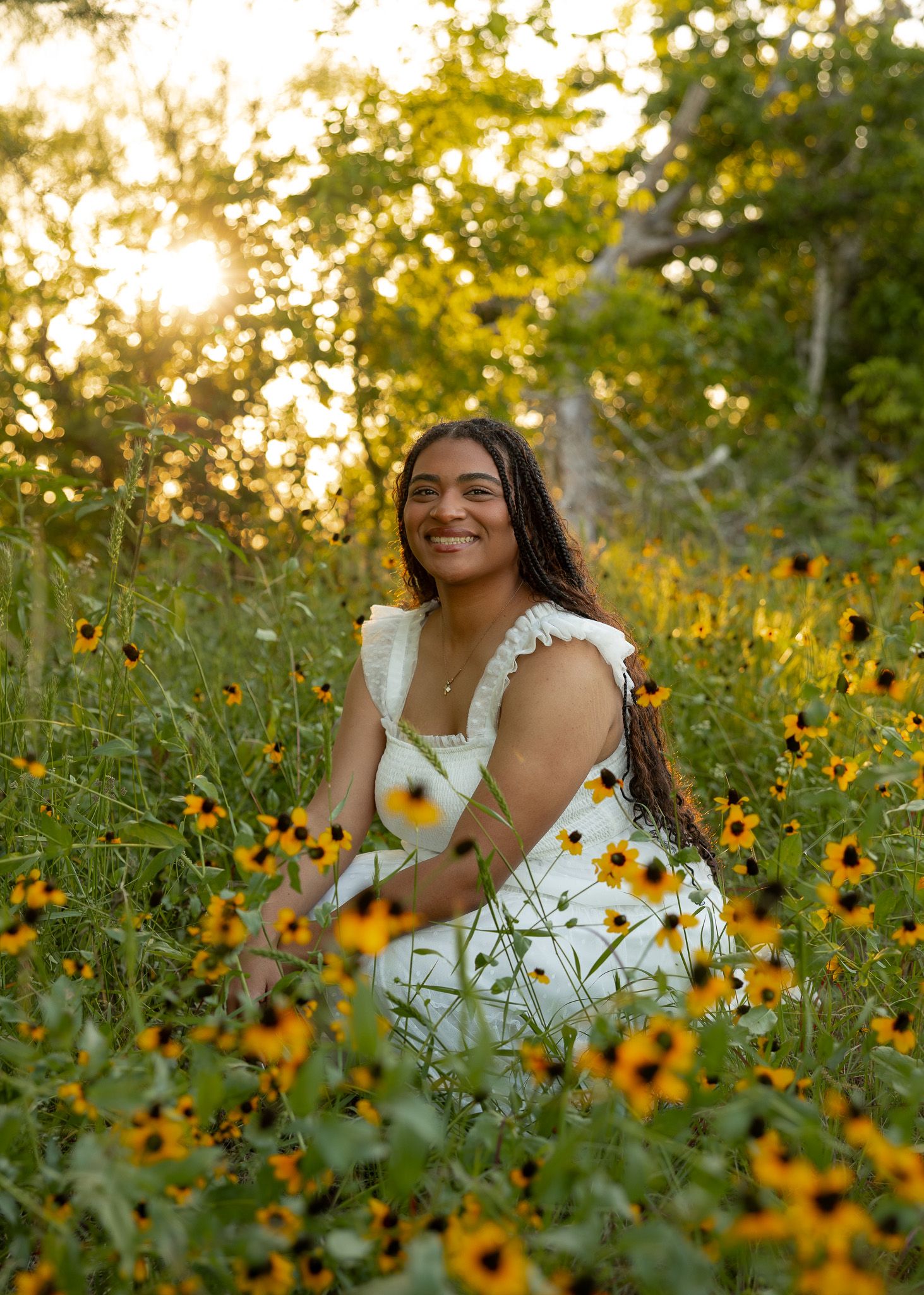 Young woman in white dress sitting among yellow black-eyed Susan wildflowers at golden hour sunset.