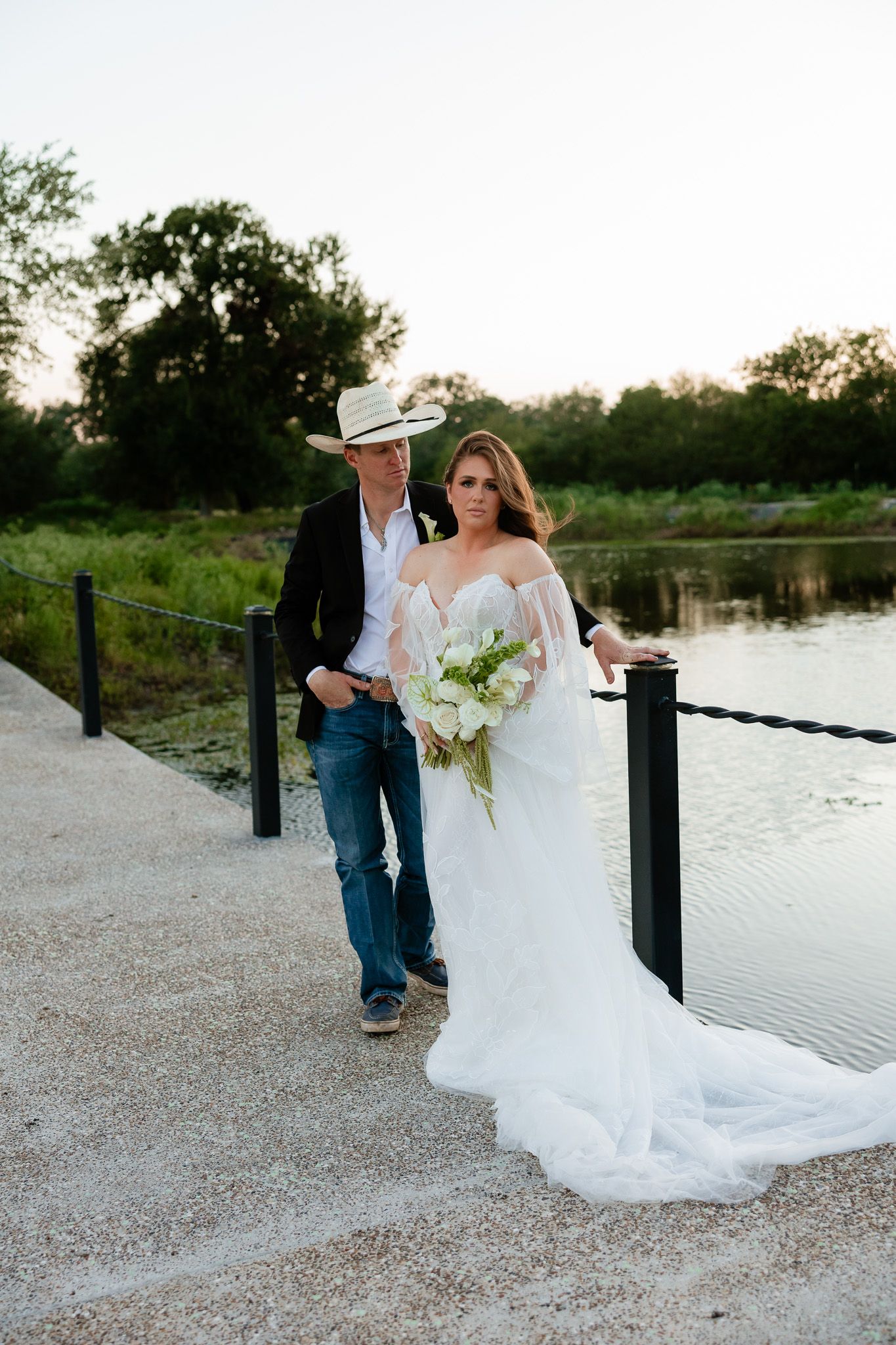 Bride in white gown holding bouquet stands with groom in cowboy hat and jeans on waterfront dock at sunset.