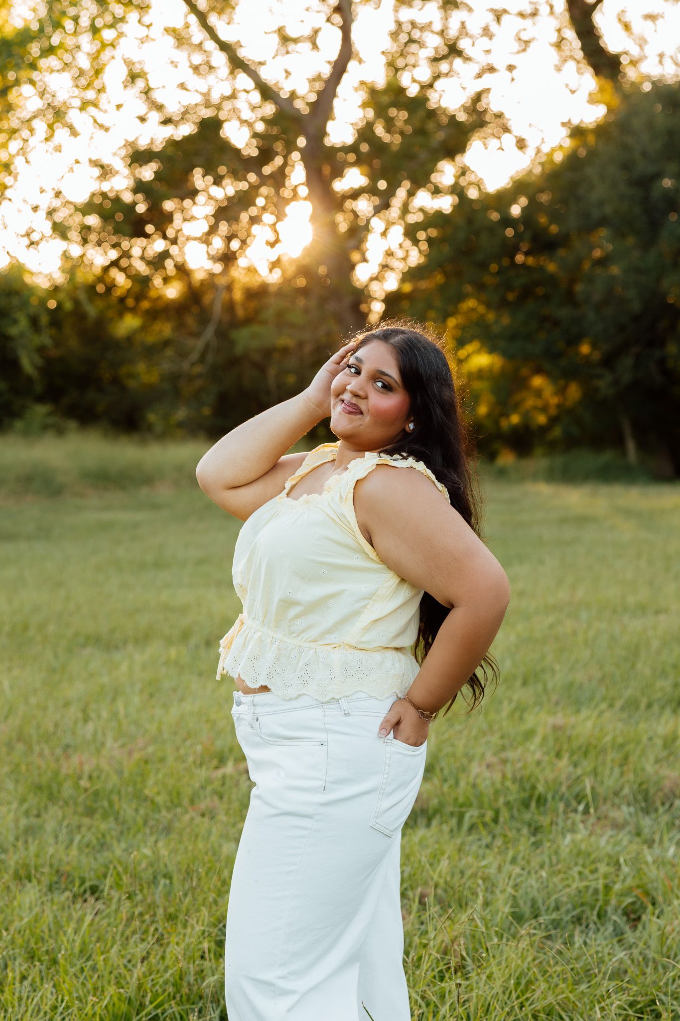 Young woman in yellow top and white pants posing outdoors at golden hour with lush green trees behind her.
