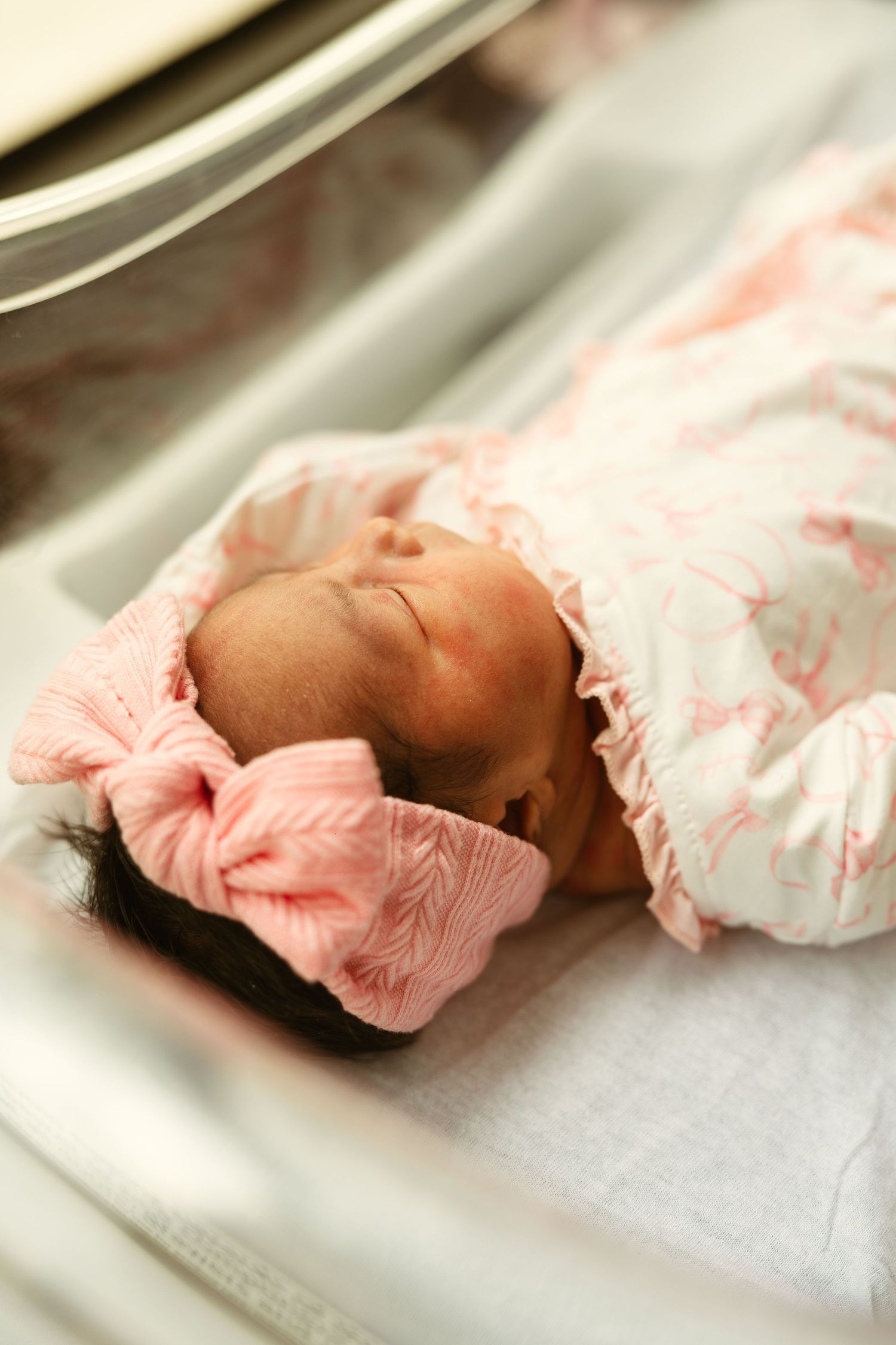 Newborn baby girl sleeping peacefully in hospital bassinet, wearing pink floral swaddle and matching bow headband.