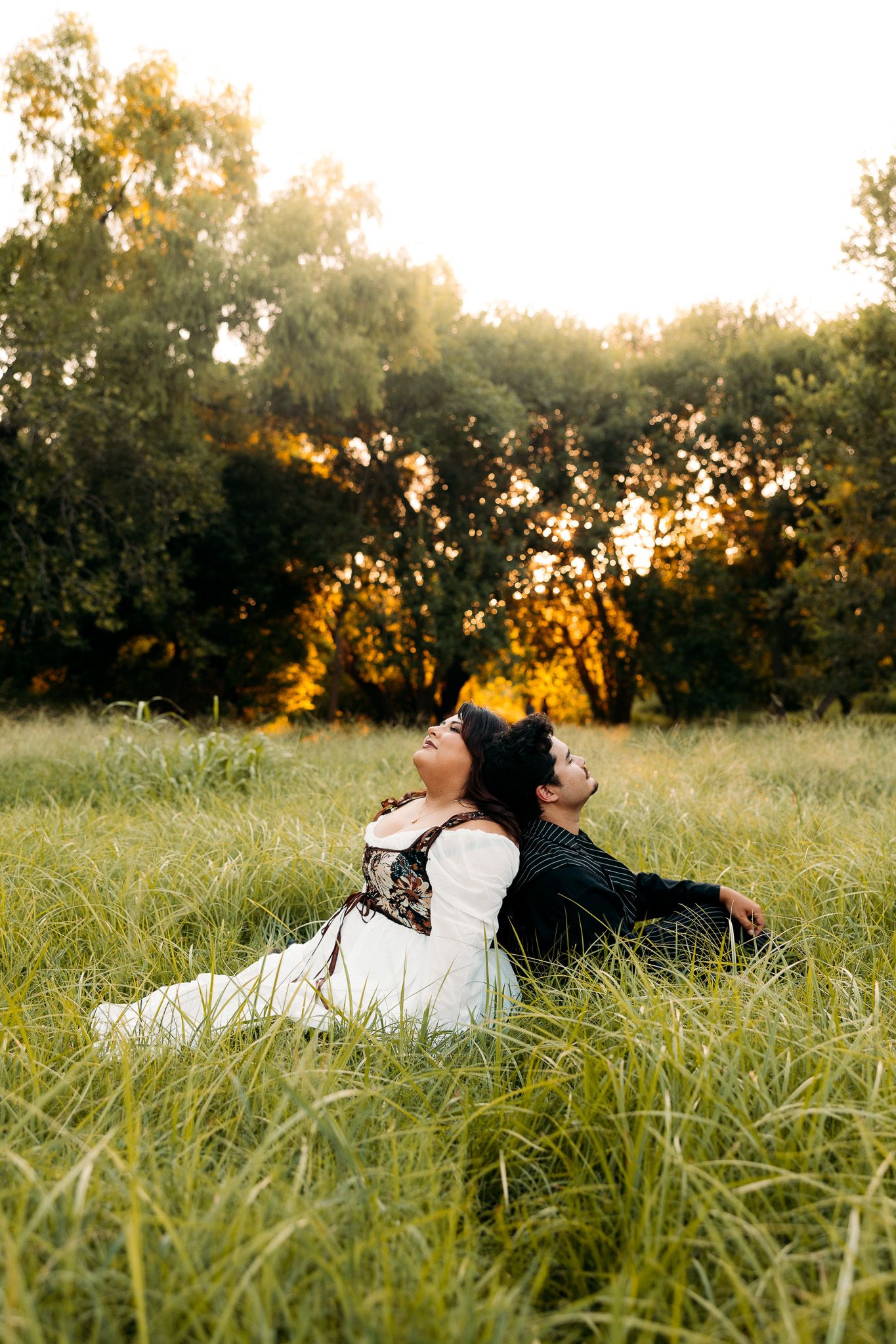 Two women in elegant dresses sit back-to-back in a lush green field at golden sunset.