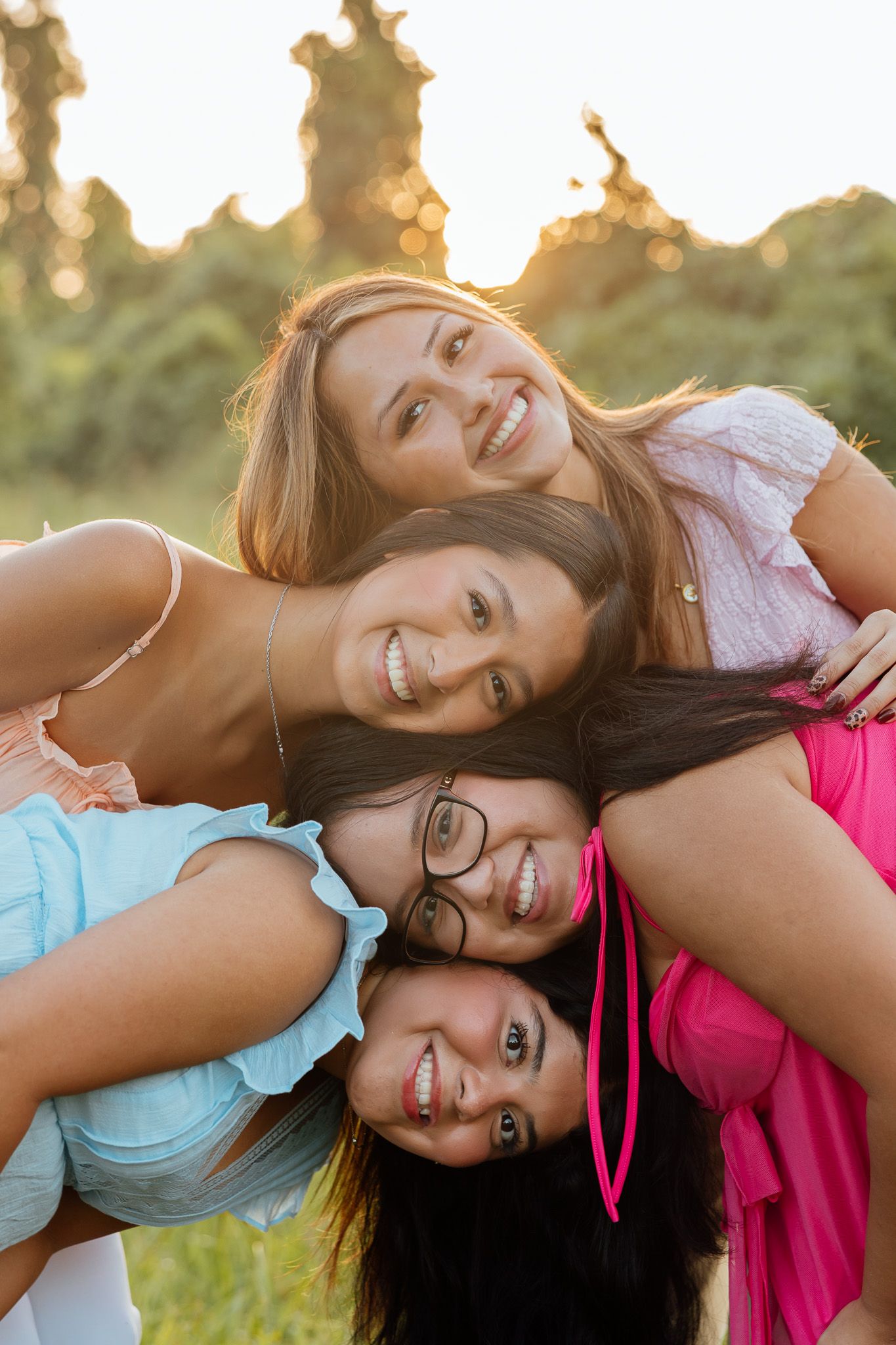 Four smiling young women huddle together outdoors in golden sunset light, posing playfully for a group photo.