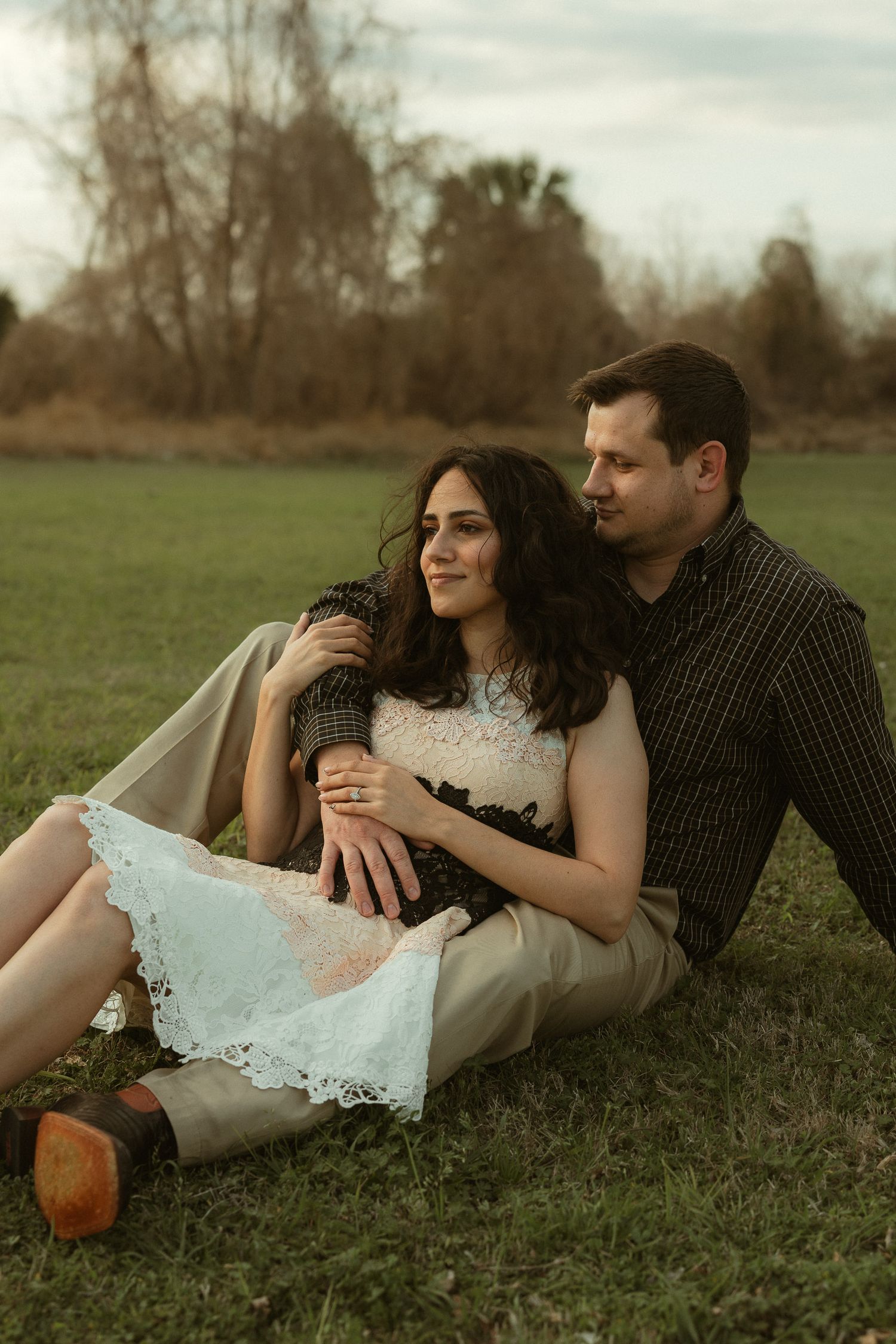 A couple sits together on grass, the woman in a white lace dress leaning against a man in a dark plaid shirt.