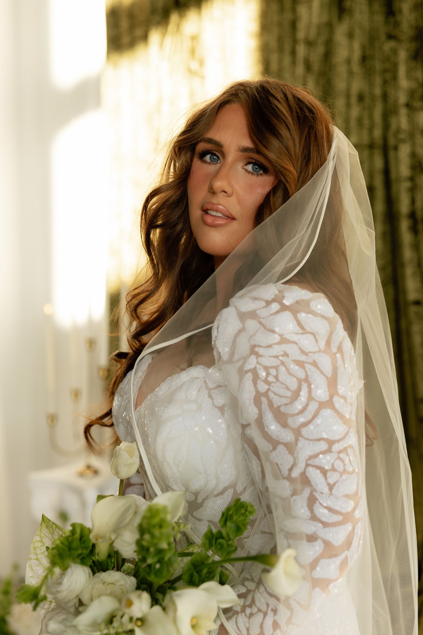 Bride in white lace gown with veil holding green and white bouquet, posing in elegant sunlit room.