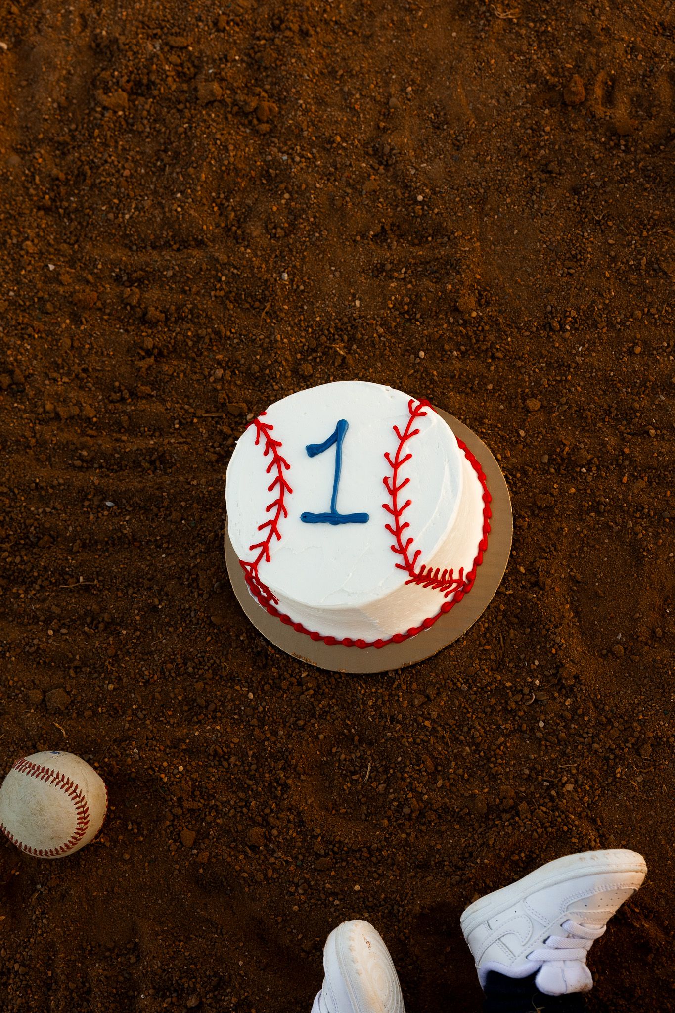 Baseball-themed first birthday cake with red stitching and blue number 1, placed on a dirt field next to a baseball.