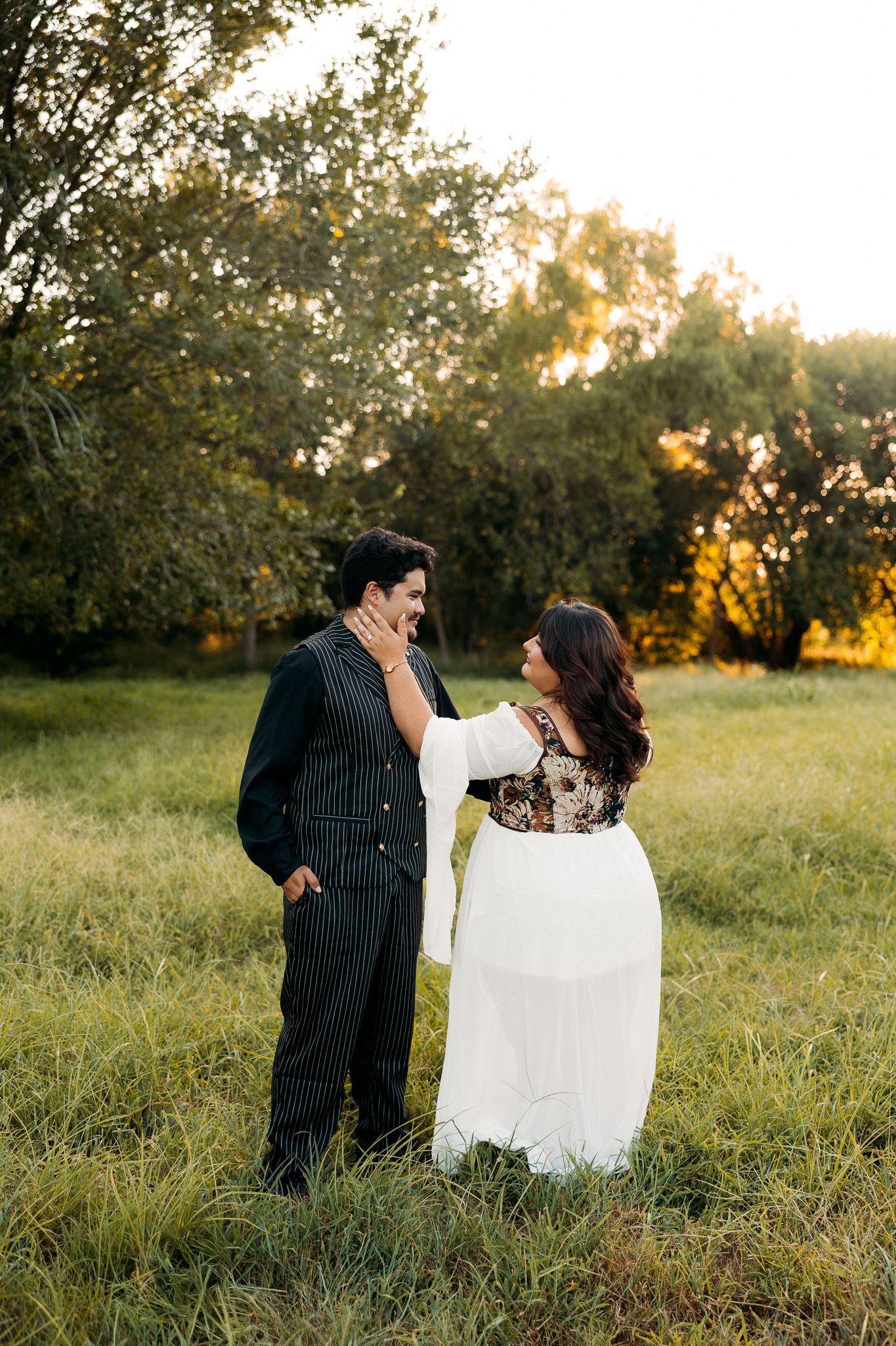 Couple in elegant attire standing in a sunlit green field, woman in white dress touching man's face tenderly.