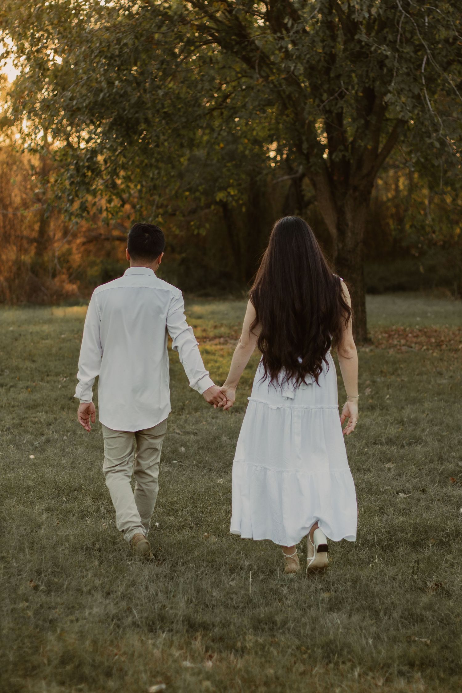 A couple in matching white outfits strolls through a grassy field at sunset.