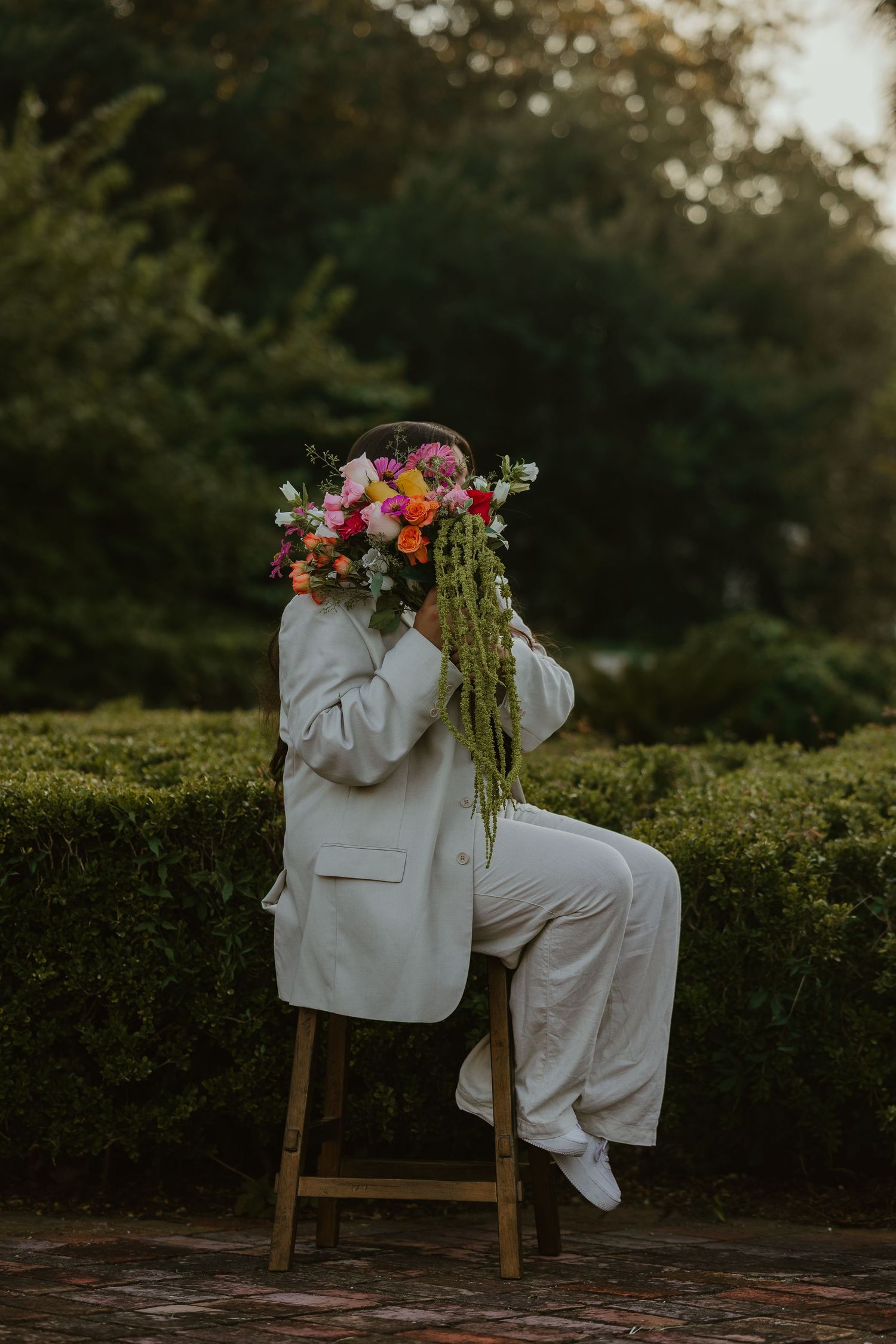 White-suited figure seated on wooden chair with colorful floral arrangement against garden backdrop.