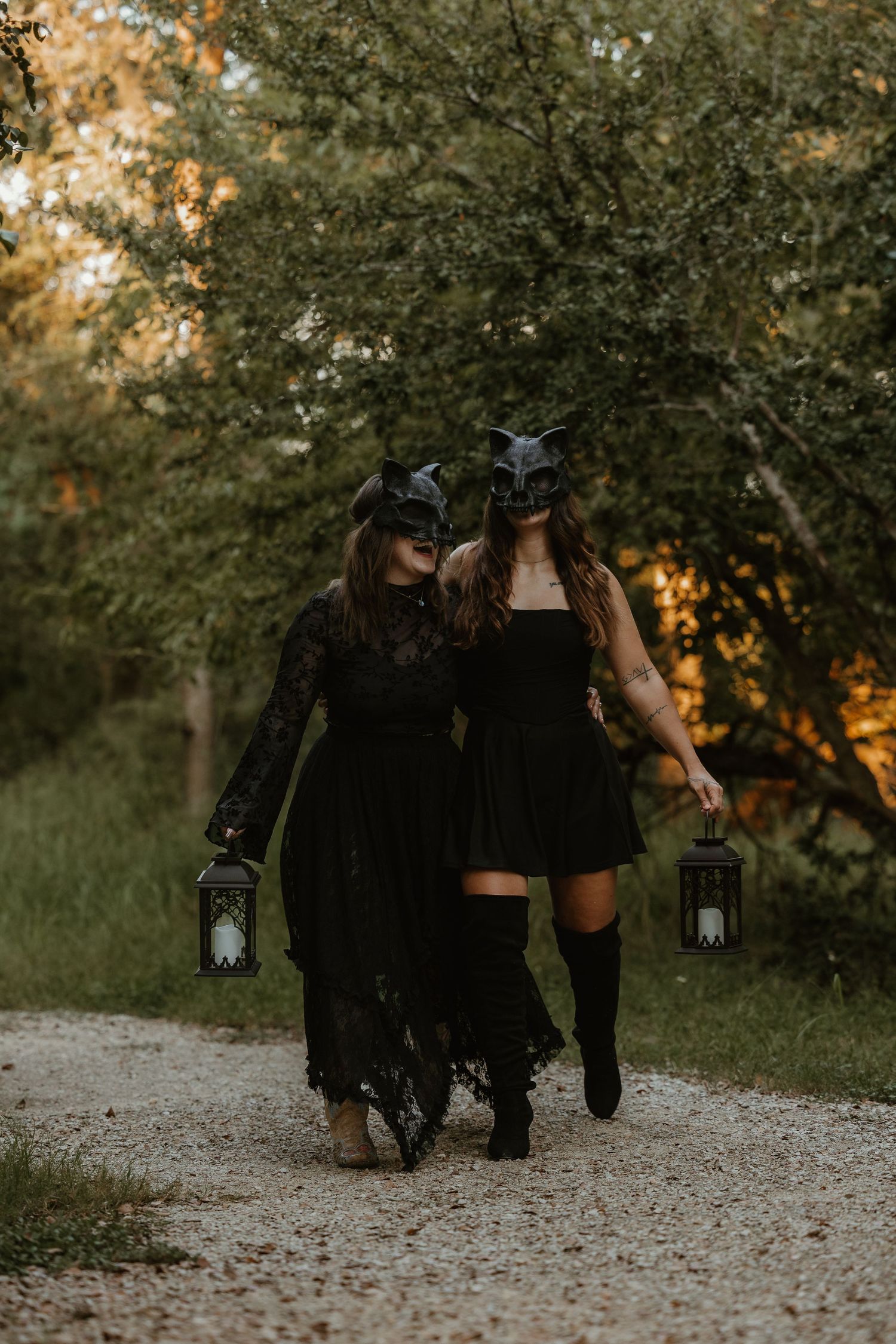 Two figures in black dresses and masks walking down a path carrying lanterns at dusk.