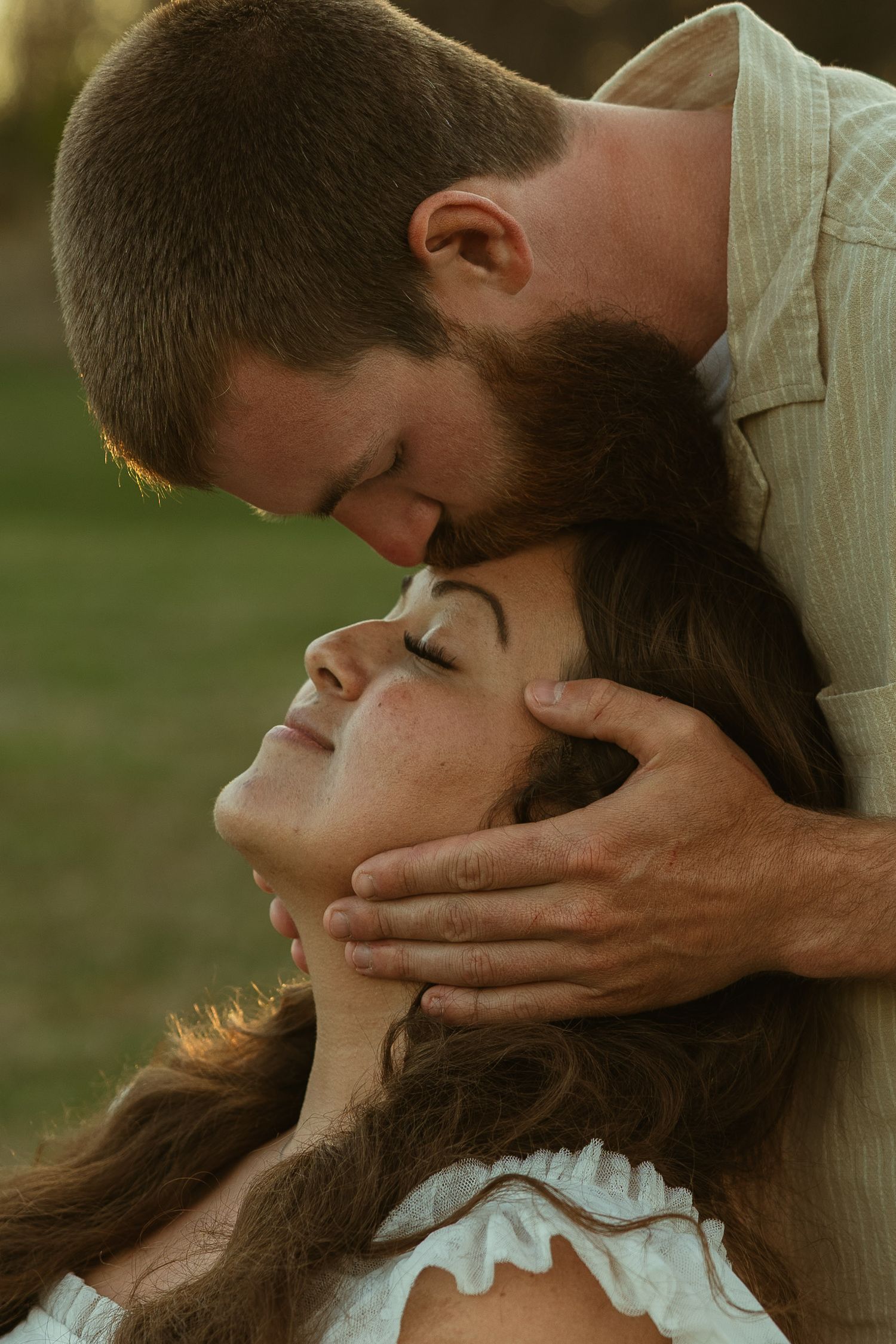 A bearded man tenderly kisses a woman's forehead while cradling her face in his hands outdoors.
