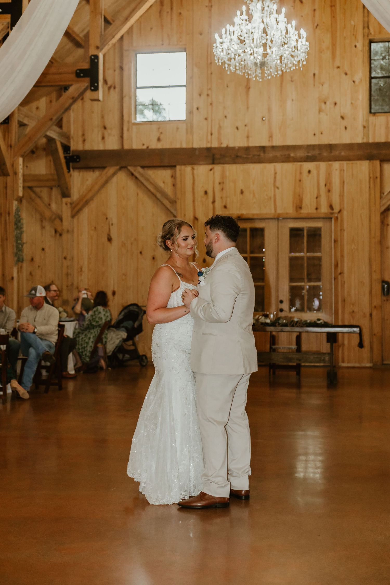 A couple shares their first dance in an elegant barn reception venue with wooden walls and chandeliers.